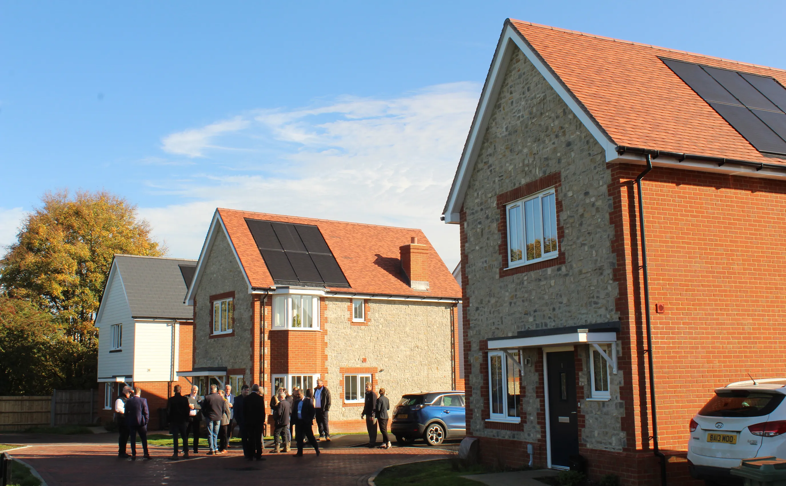 People stand outside a row of newly built brick and stone houses with rooftop solar panels, under a clear blue sky. Parked cars line the driveway, and the setting suggests a sunny community event focused on sustainable residential development.