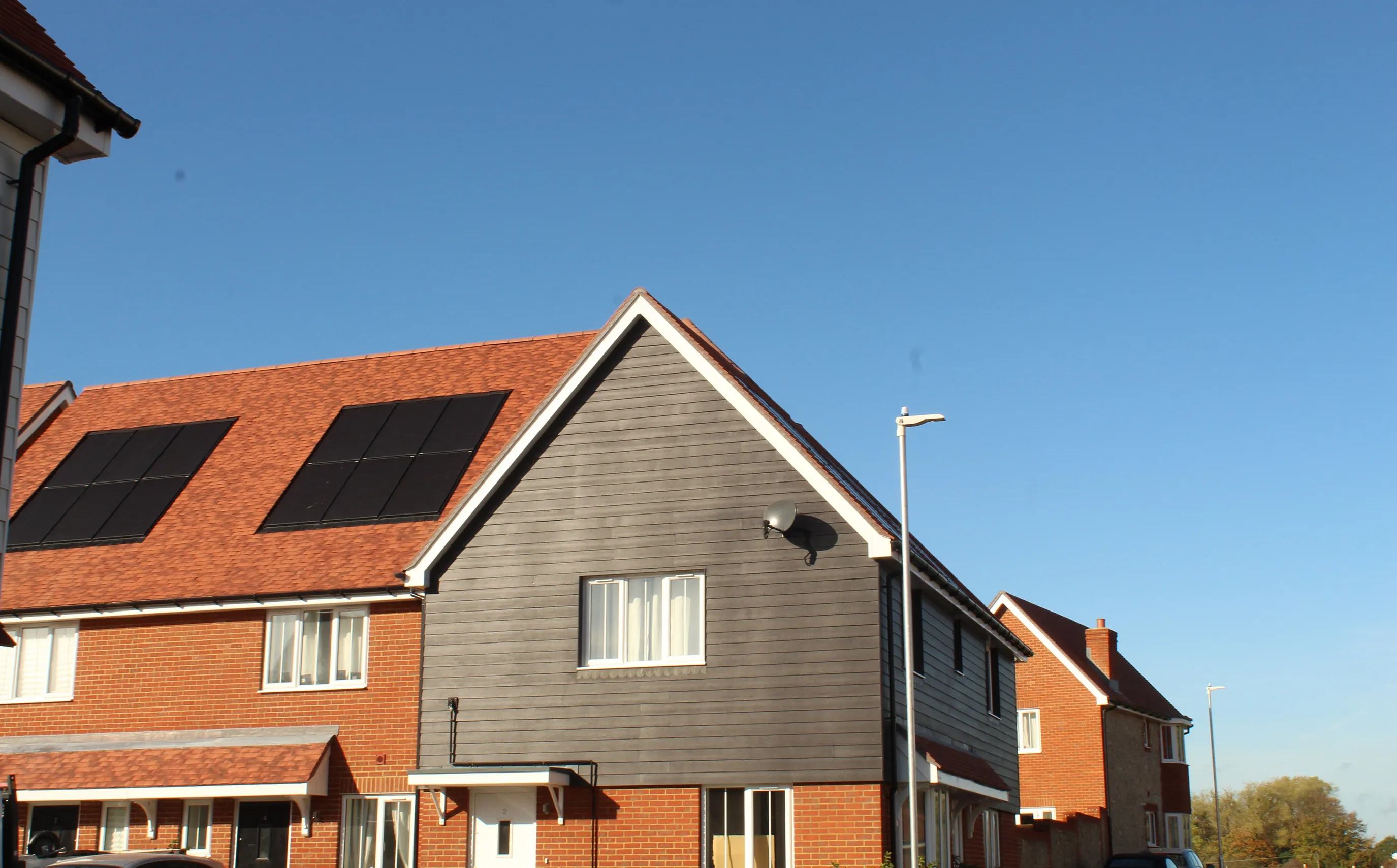 A row of contemporary homes featuring red-tiled roofs and a mix of brick and timber façades. The house in the foreground is fitted with sleek solar panels, emphasizing a commitment to renewable energy. A streetlamp stands nearby under a vivid blue sky, highlighting the neighbourhood’s clean, modern aesthetic.