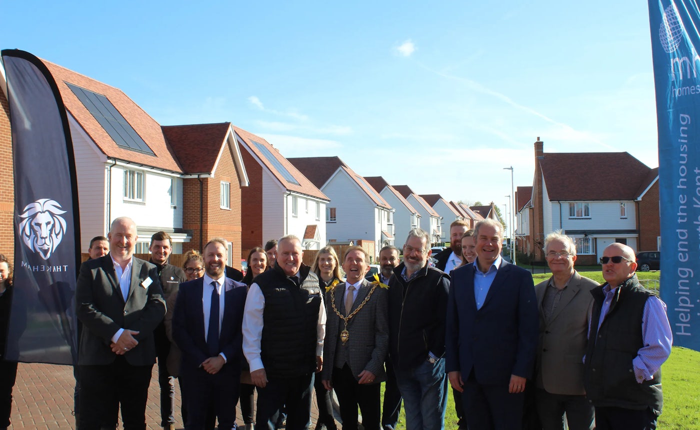 A group of people in formal and semi-formal clothing stand in front of a row of modern houses with red-tiled roofs. Two large banners are displayed: one with the logo and text 'THAKEHAM,' and the other reading 'Helping end the housing.' The scene takes place on a sunny day, suggesting a community event or housing-related initiative.