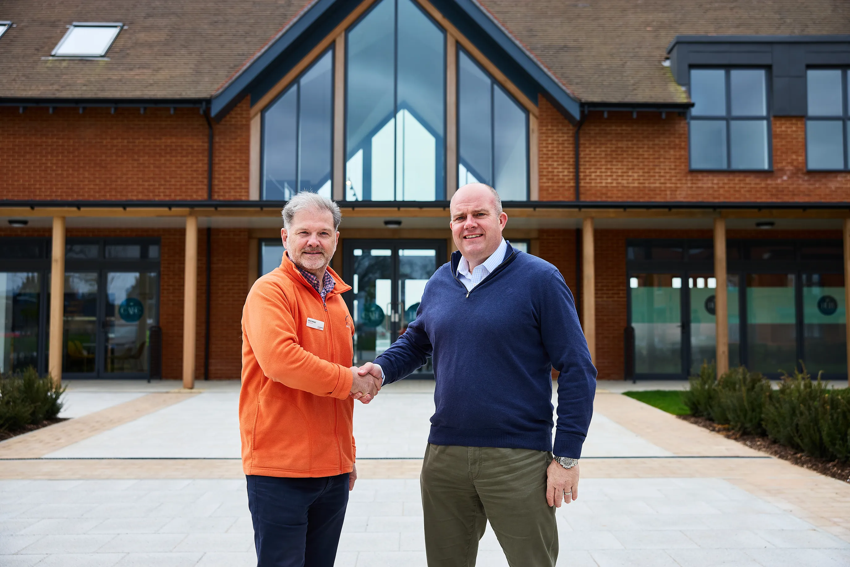 Two individuals shake hands outside a modern building with large windows and a red-brick exterior. One wears an orange jacket and the other a blue sweater, suggesting a casual yet professional interaction, possibly marking an agreement or partnership."