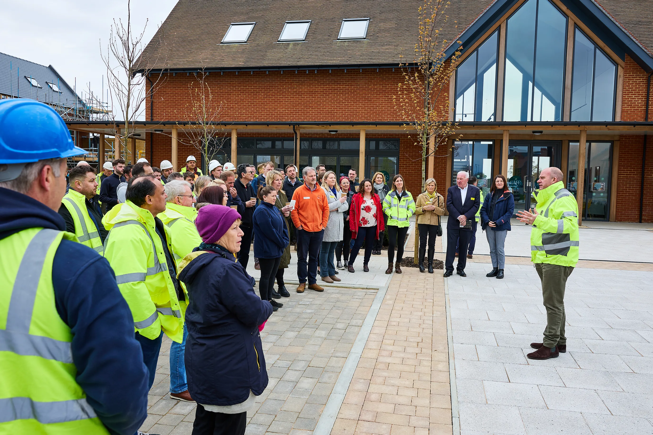 A group of people, including several in high-visibility jackets and construction helmets, gather outside a modern brick building with large glass windows. One individual appears to be addressing the group, suggesting a site tour, briefing, or milestone event at a construction or recently completed development site.