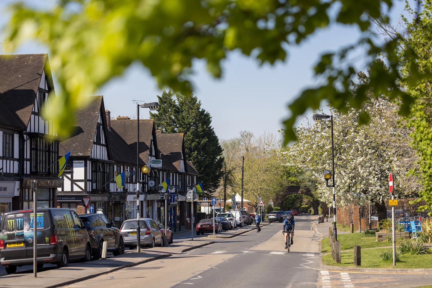 A local street flanked by charming half-timbered buildings with white plaster and dark wooden beams, likely from a historic English village. Cars are parked along one side, while a lone cyclist rides through the quiet scene. Leafy trees line the sidewalk, their fresh spring foliage hinting at a mild, pleasant day. The overall atmosphere is serene and nostalgic, evoking a sense of timelessness and community.