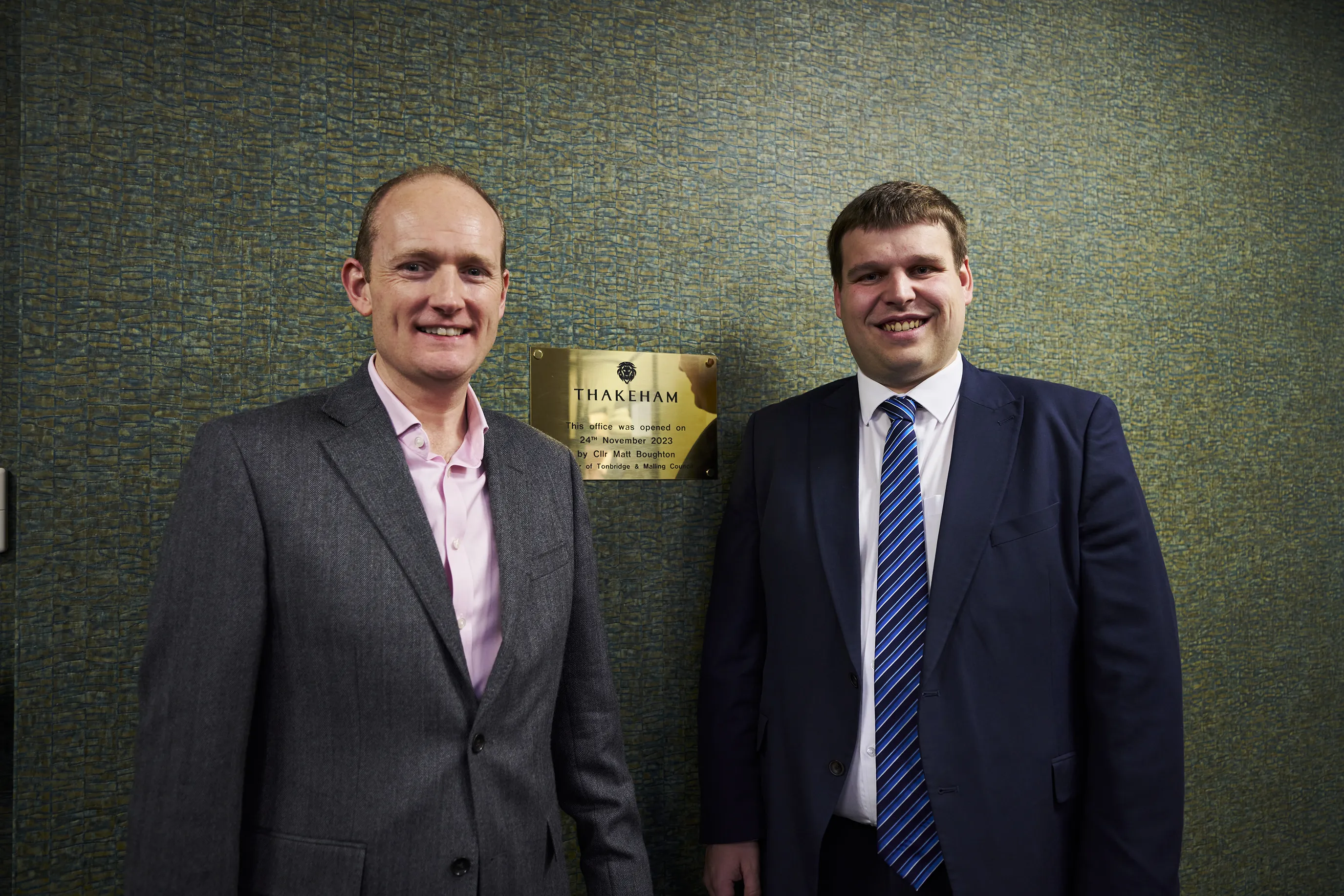 Two people stand in front of a light brick wall featuring a commemorative plaque. The plaque reads: “Thakeham – Building a legacy. This plaque was unveiled by Rt Hon Michael Gove MP, Secretary of State for Levelling Up, Housing and Communities, on 17th February 2022.” The individuals are dressed formally and smiling, with the scene suggesting a ceremonial or official event.