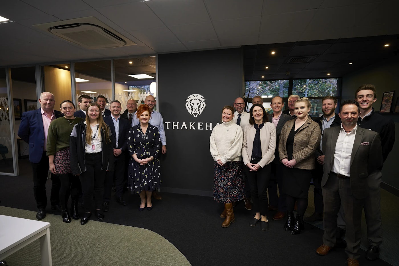 A group of professionally dressed people pose in front of a wall displaying the Thakeham logo, which features a stylized lion’s head above bold lettering. The setting appears to be a modern office environment with glass walls and natural light. The group stands close together, suggesting a team event or corporate gathering.