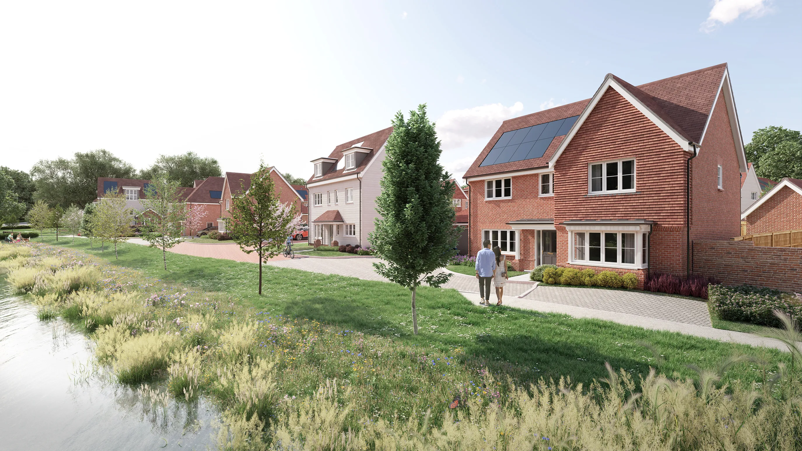 Two people stroll along a tree-lined path in a sunny, eco-conscious neighbourhood. Brick houses with solar panels and manicured front gardens line one side, while wildflowers and a reflective body of water stretch alongside the other, creating a calm, community-focused setting.