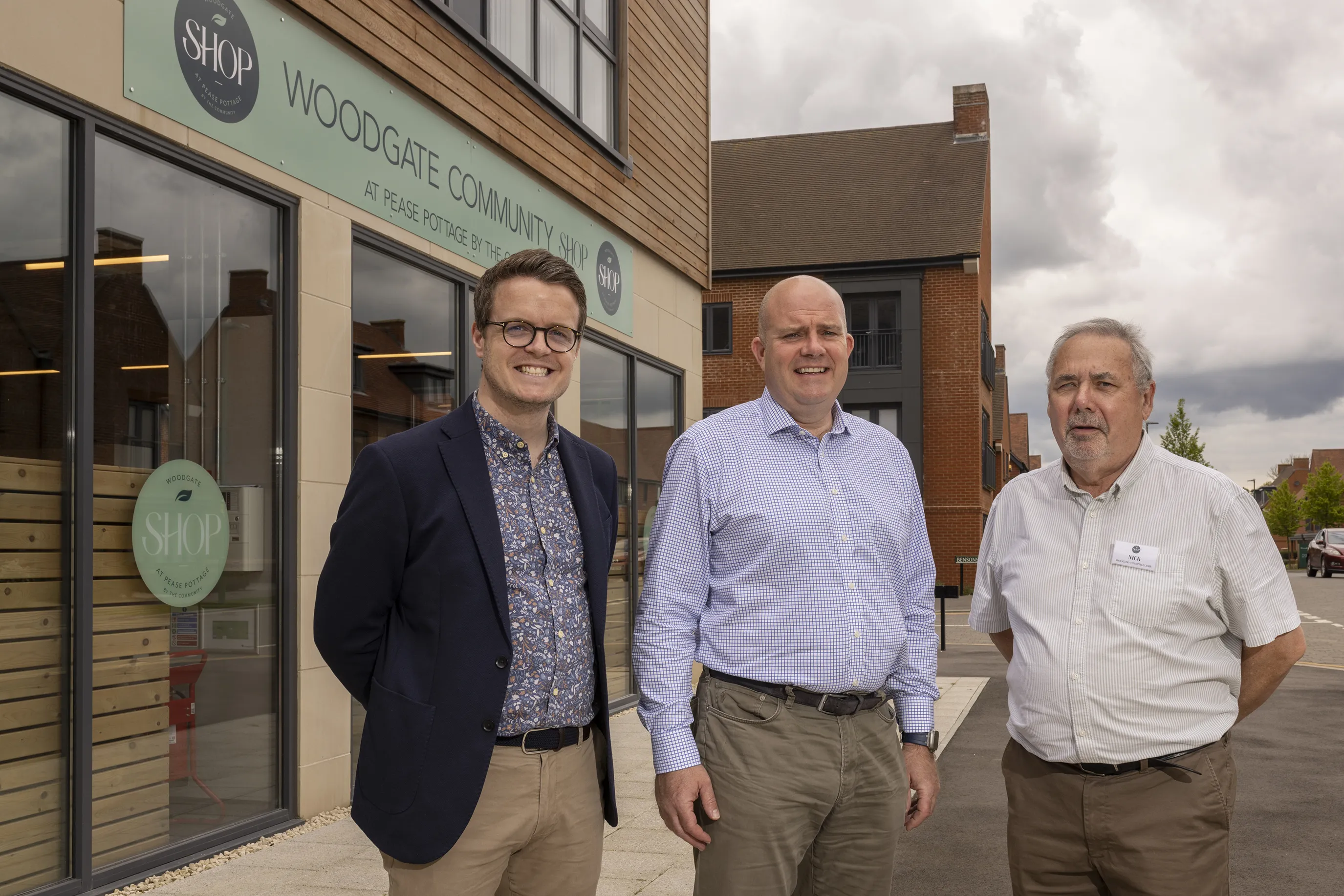 Three people stand smiling outside a brick building with a sign reading “Woodgate Community Shop at Pease Pottage.” The group is dressed in smart-casual clothing, suggesting a celebratory moment, possibly marking the shop’s opening. Modern homes and a cloudy sky appear in the background.