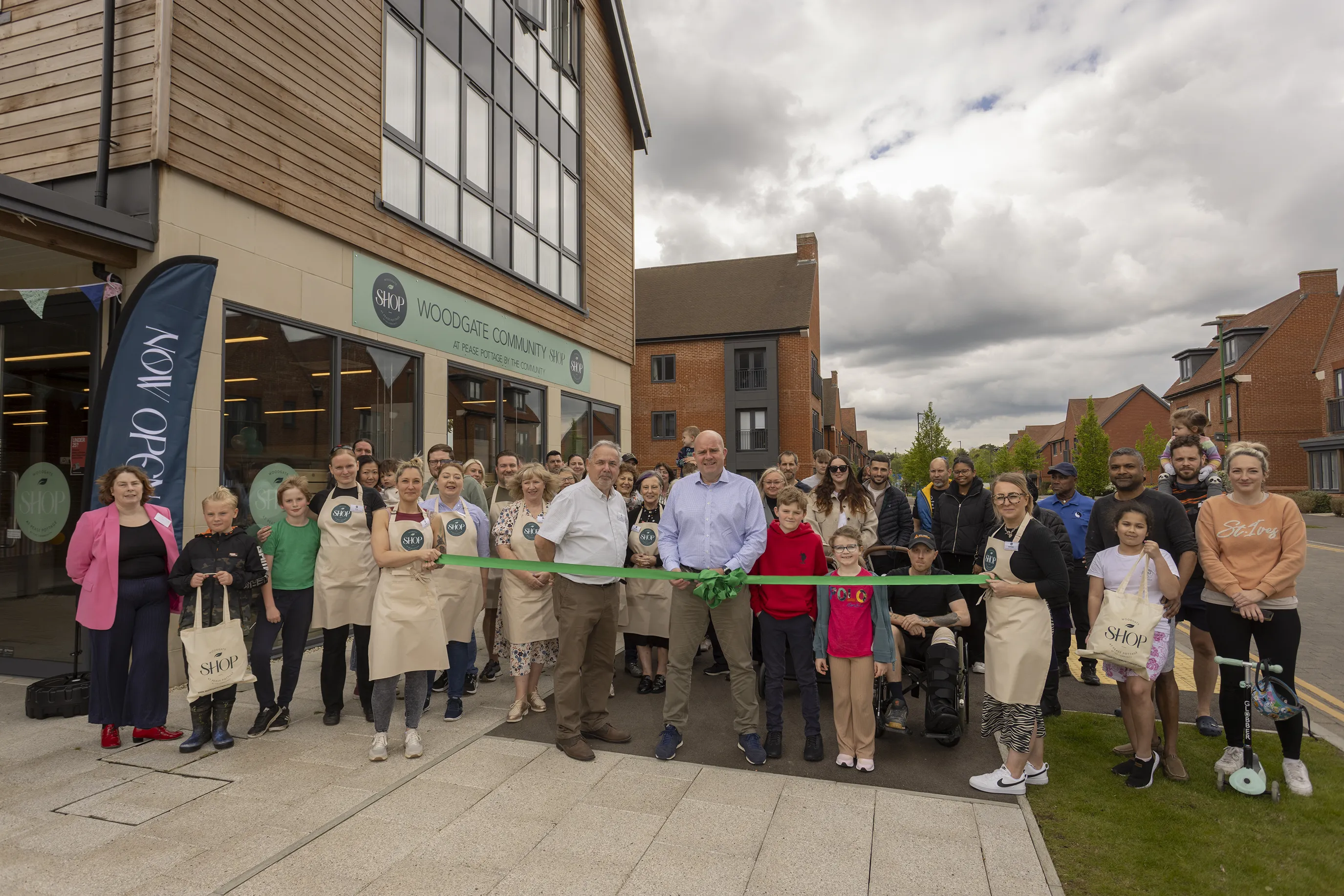 A group of people pose smiling in front of a newly opened building marked “WOODGATE COMMUNITY SHOP” as part of a ribbon-cutting ceremony. Several wear branded aprons, and one person holds ceremonial scissors near a green ribbon. A banner to the side reads “NOW OPEN,” emphasizing the celebratory tone of the event.