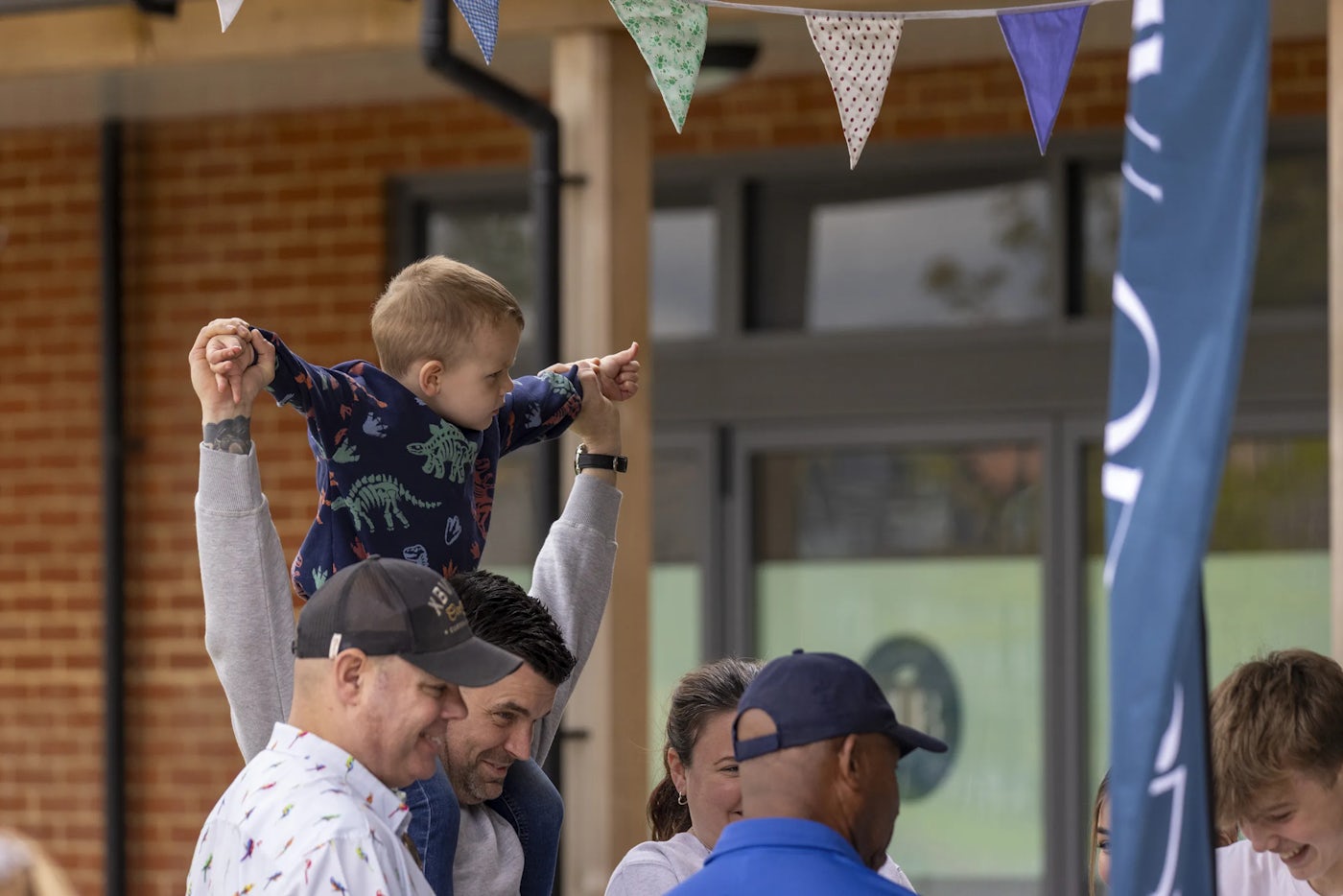 A group of people gather outdoors beneath colorful triangular bunting, creating a festive atmosphere. One person lifts a child into the air as the child points excitedly into the distance. A brick wall and windowed building form the backdrop, suggesting a cheerful community event.