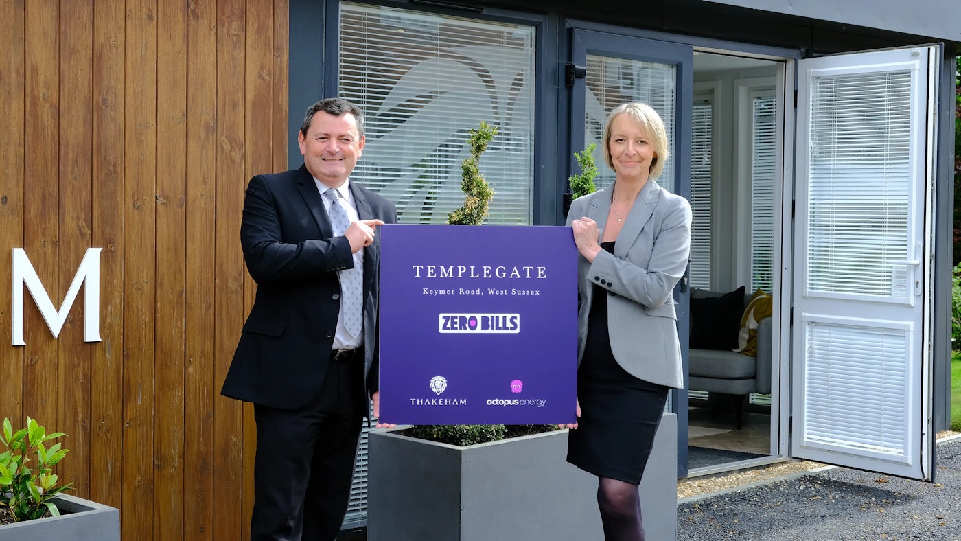 Two people standing outside a modern wood-clad building, holding a sign reading 'TEMPLEGATE, Keymer Road, West Sussex, ZERO BILLS, THAKEHAM, Octopus Energy,' with large planters and a visible interior lounge—suggesting an eco-conscious housing initiative.