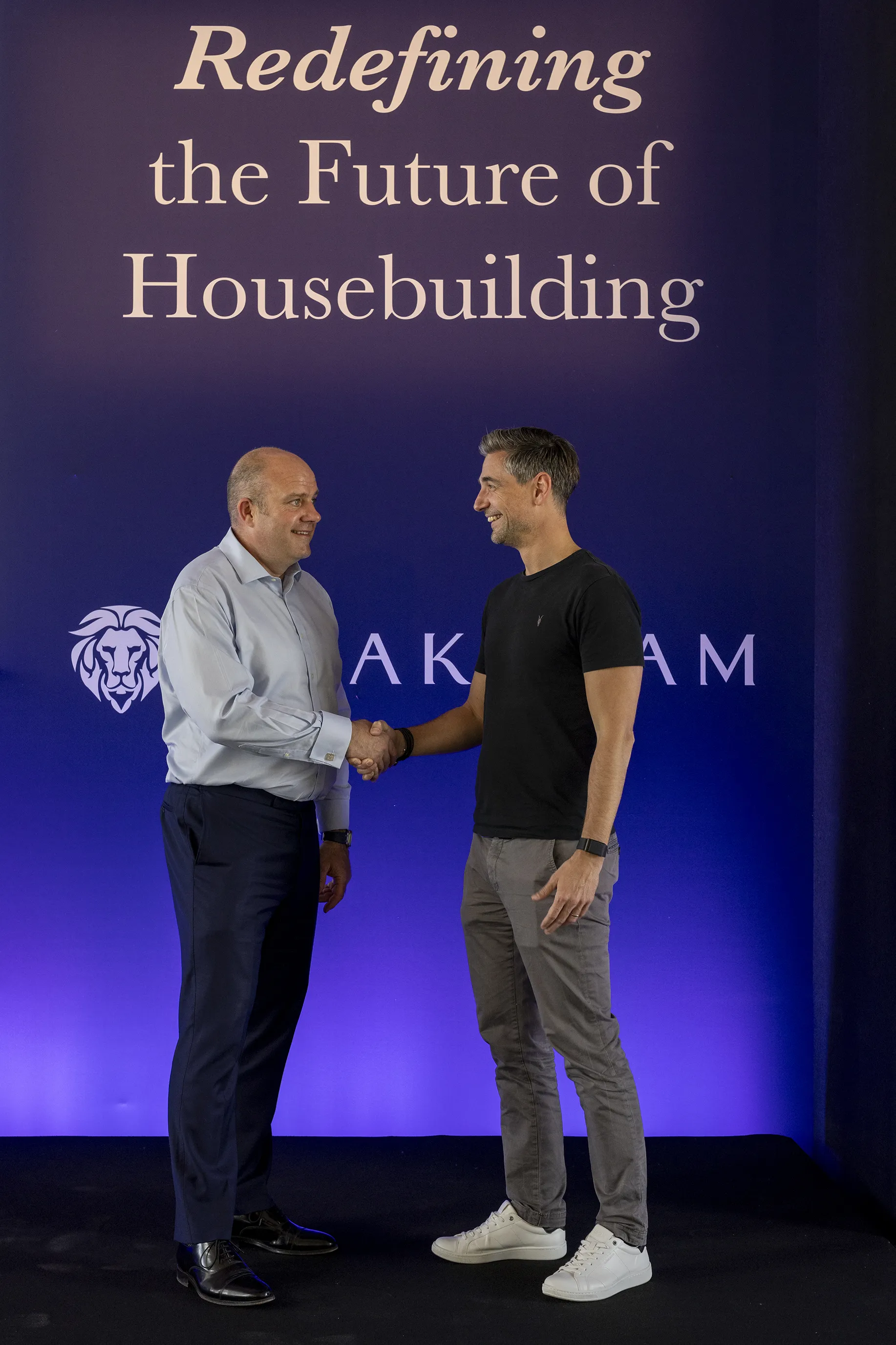 Two people shaking hands in front of a dark blue-to-purple gradient banner reading 'Redefining the Future of Housebuilding,' with a stylized lion’s head logo and the word 'THAKEHAM' below—suggesting a formal event or partnership in the housing sector.