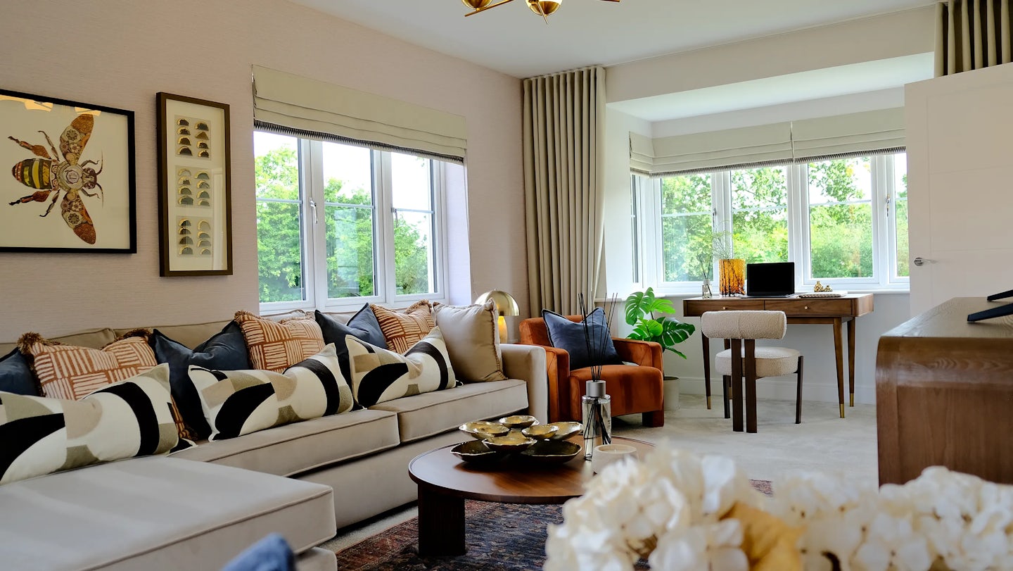 Stylish living room with a large L-shaped sofa adorned with patterned cushions, a round coffee table, and a desk setup with a laptop in the corner. A framed bee illustration hangs above the sofa, while natural light and greenery outside the window enhance the cozy, modern ambiance.