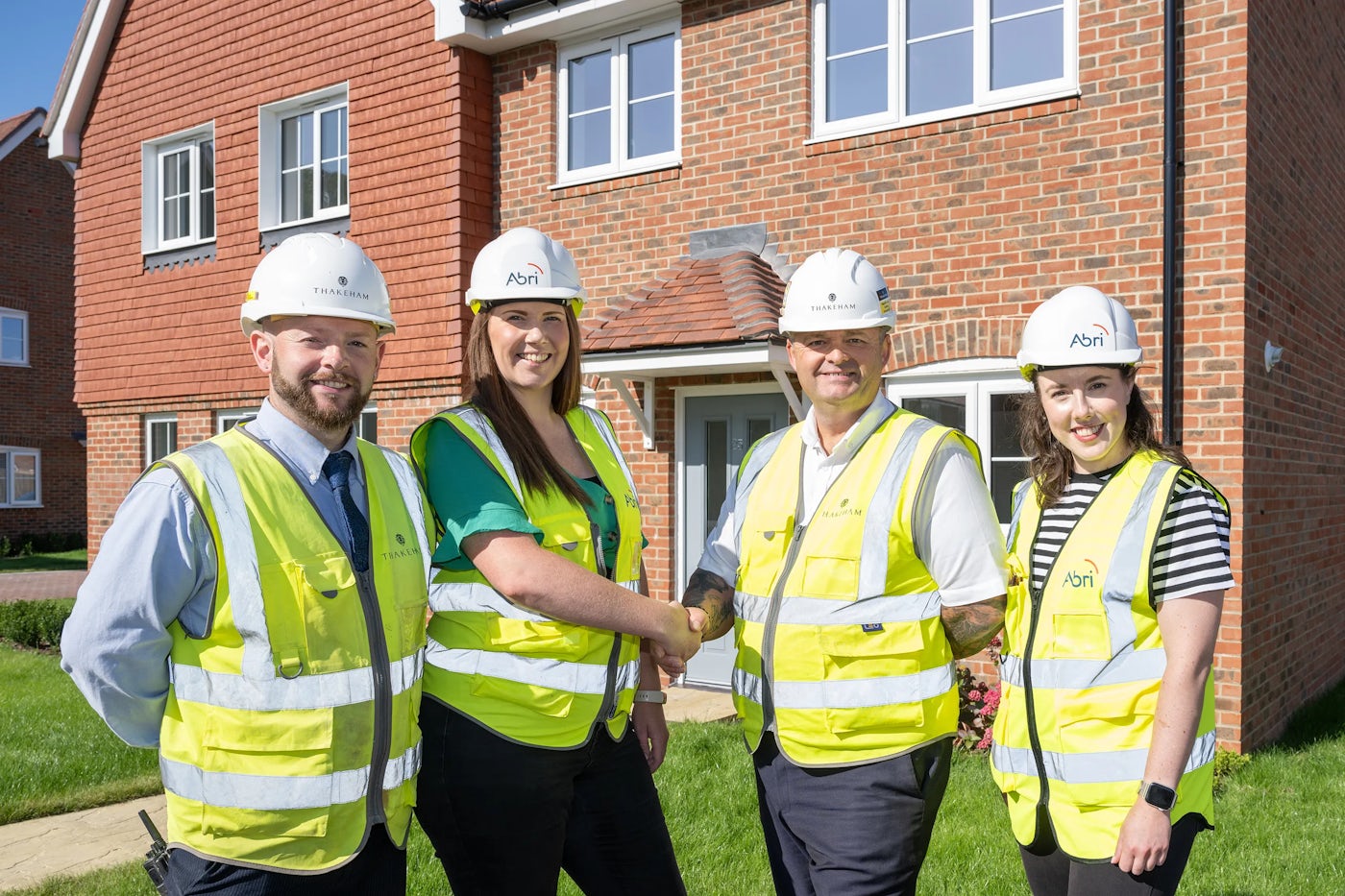 Four people in high-visibility vests and Abri-branded hard hats standing outside a newly built red-brick home, with two shaking hands—signaling project completion or a new partnership in housing development.