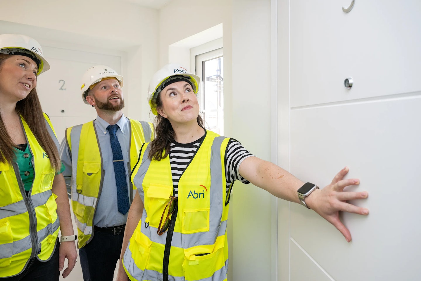 Three individuals in Abri-branded high-visibility vests and hard hats stand in a corridor outside a door marked '9,' with one person gesturing toward it—suggesting a property inspection, site handover, or housing development milestone.
