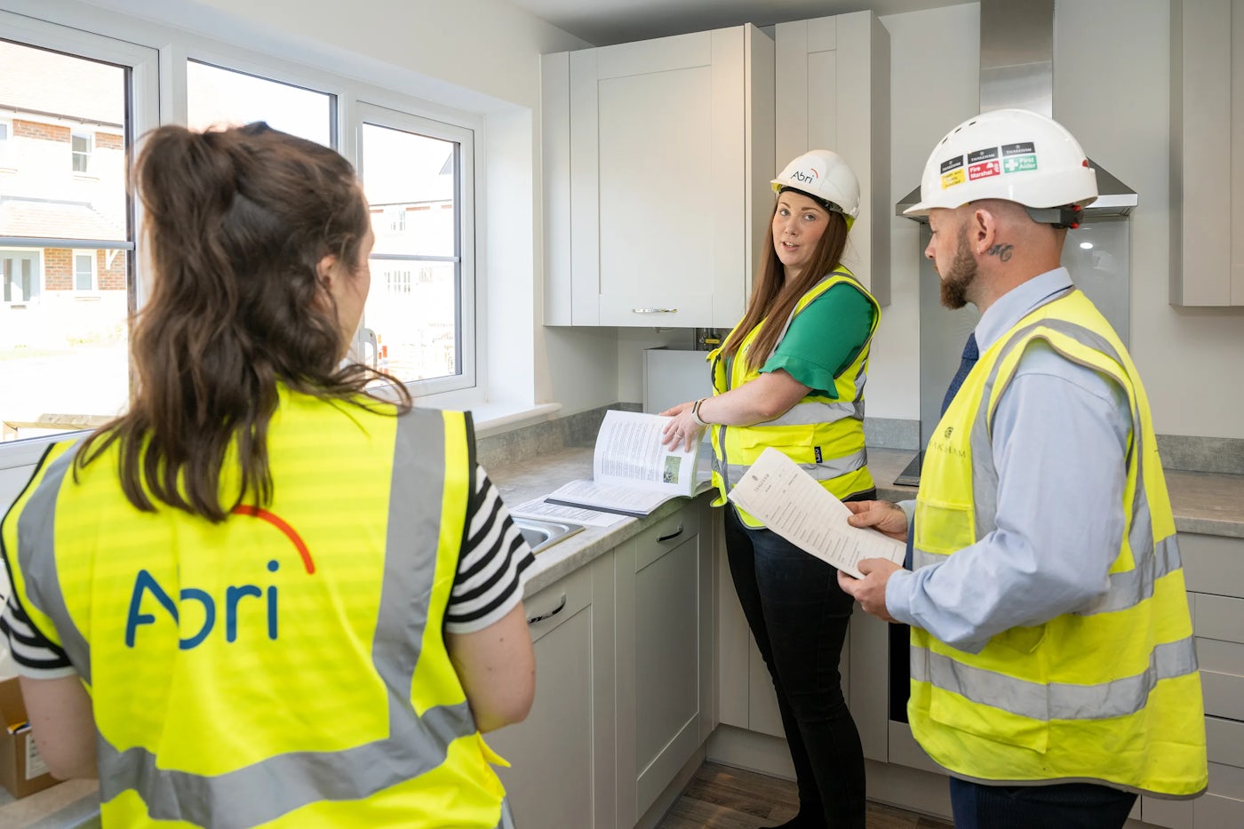 Three people wearing high-visibility vests and hard hats—one with 'Abri' on the back—stand in a newly finished kitchen, reviewing documents as part of an inspection or handover process. The setting features white cabinetry, a clean countertop, and a window providing natural light, evoking a sense of completion and readiness.