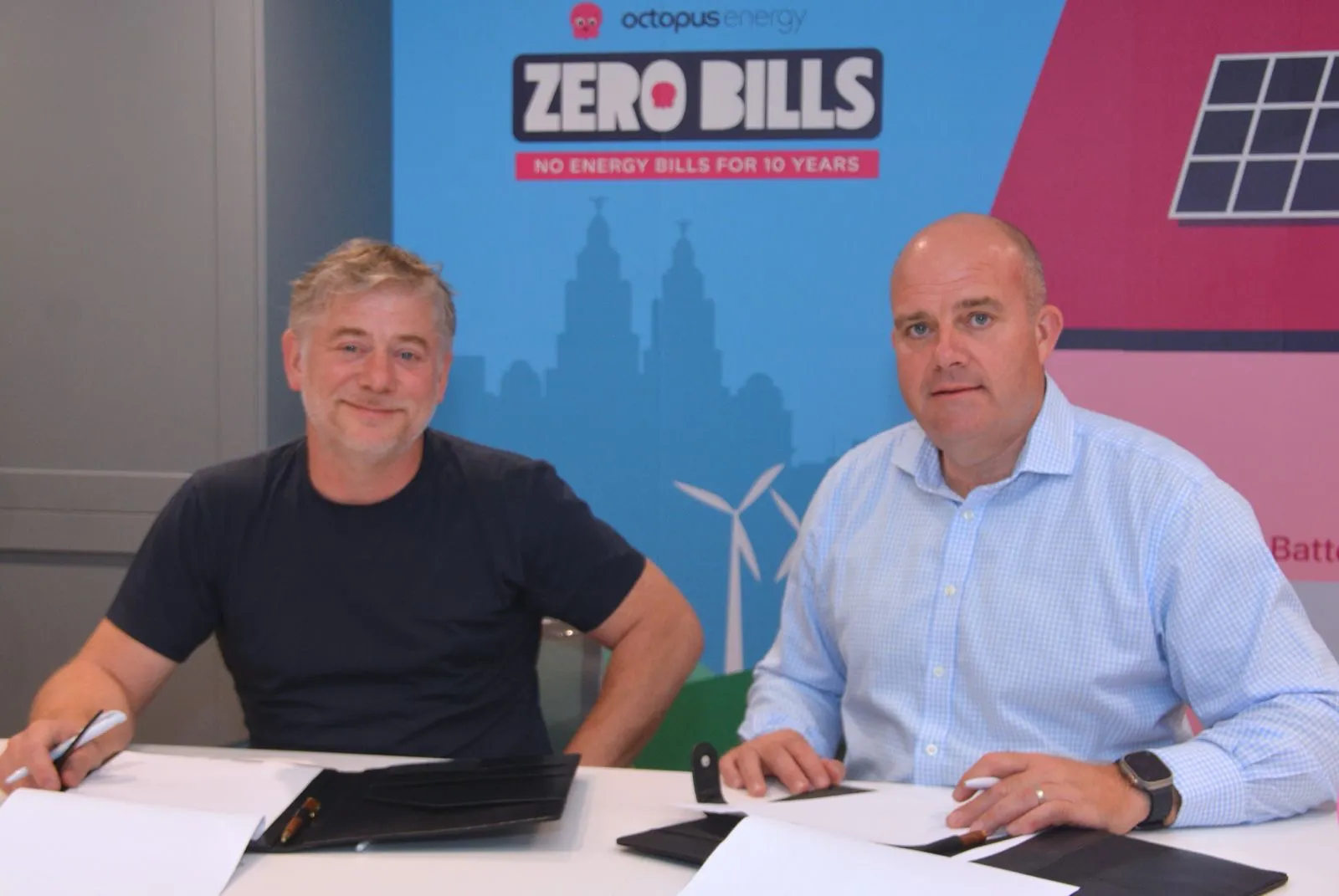 Two individuals seated at a table with pens and documents, in front of a banner that reads 'Octopus Energy ZERO BILLS NO ENERGY BILLS FOR 10 YEARS,' accompanied by silhouettes of homes, solar panels, and wind turbines—signifying a renewable energy agreement or milestone in sustainable housing.