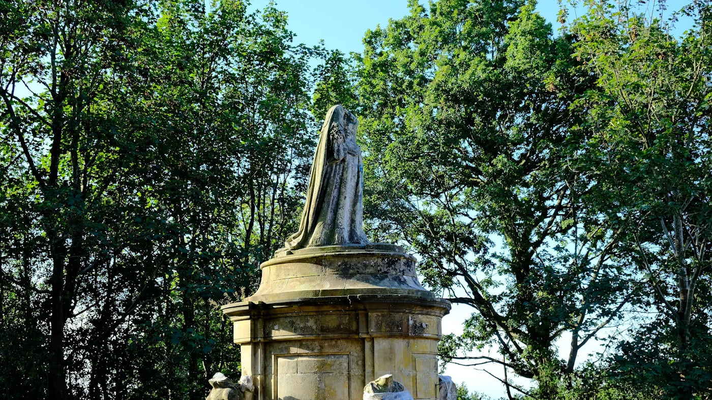 Weathered stone statue of Queen Anne atop an ornate pedestal, set against a backdrop of dense green trees—evoking a serene, timeless presence within a garden or parkland setting.