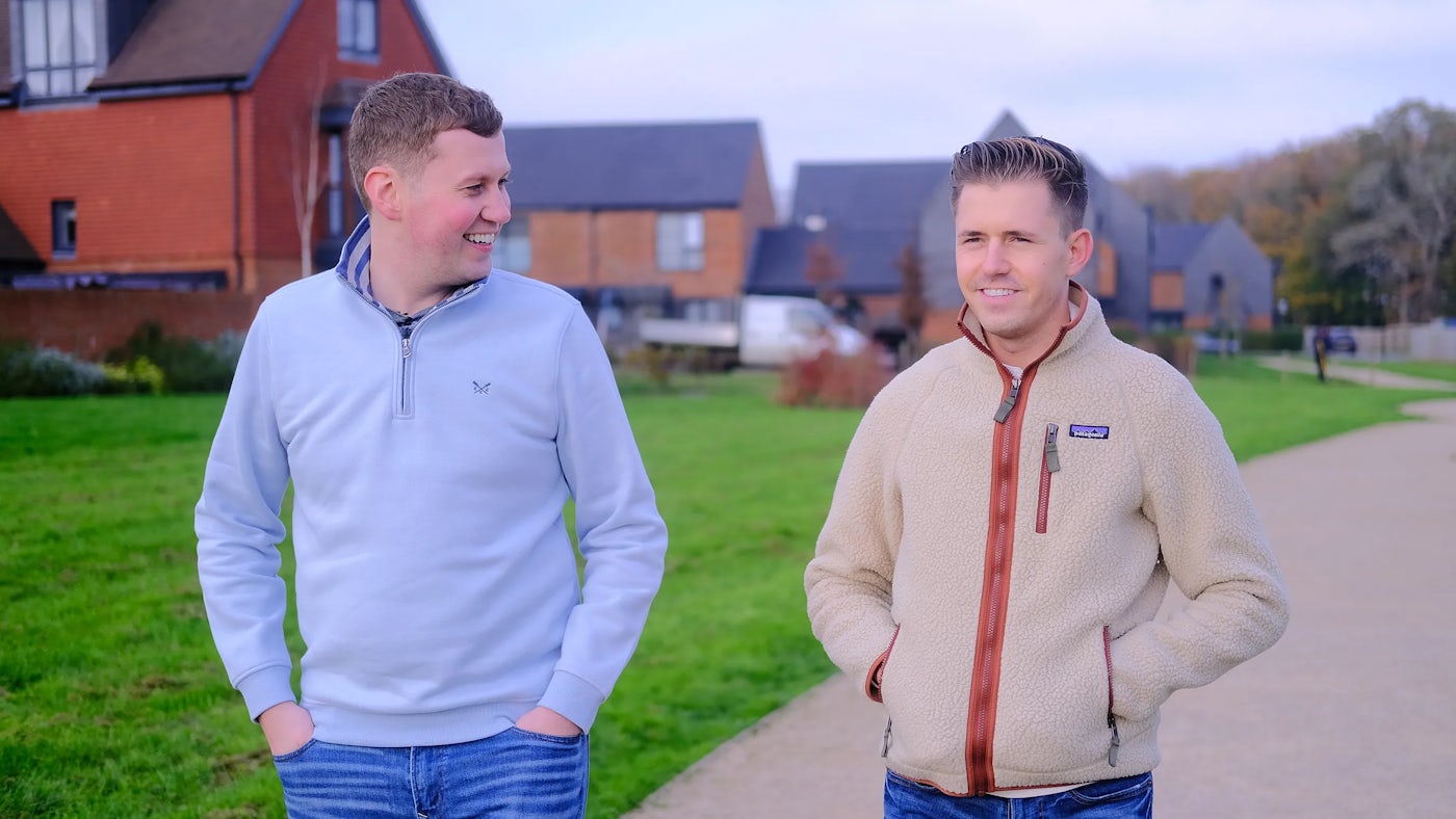 Two people stand casually in a residential area with a backdrop of modern brick homes and a small grassy space. One wears a light blue jumper, the other a beige fleece with red accents. Their relaxed posture and subtle smiles suggest a friendly chat or walk on a cool day.