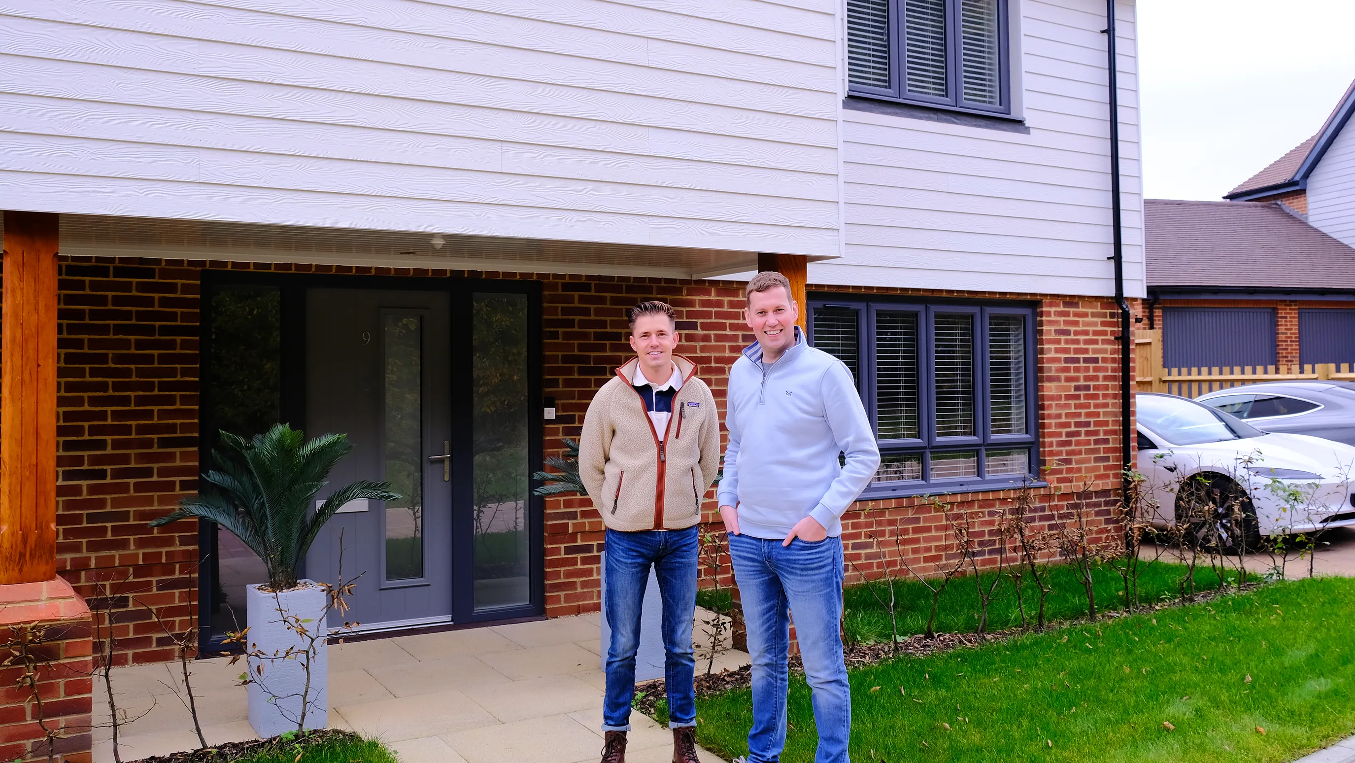 A couple stand smiling outside a modern semi-detached home with a mix of white siding and brickwork. The front door displays the number 9, and a potted plant rests near the entrance. A white car is parked in the driveway, adding to the impression of a tidy, welcoming residential setting.