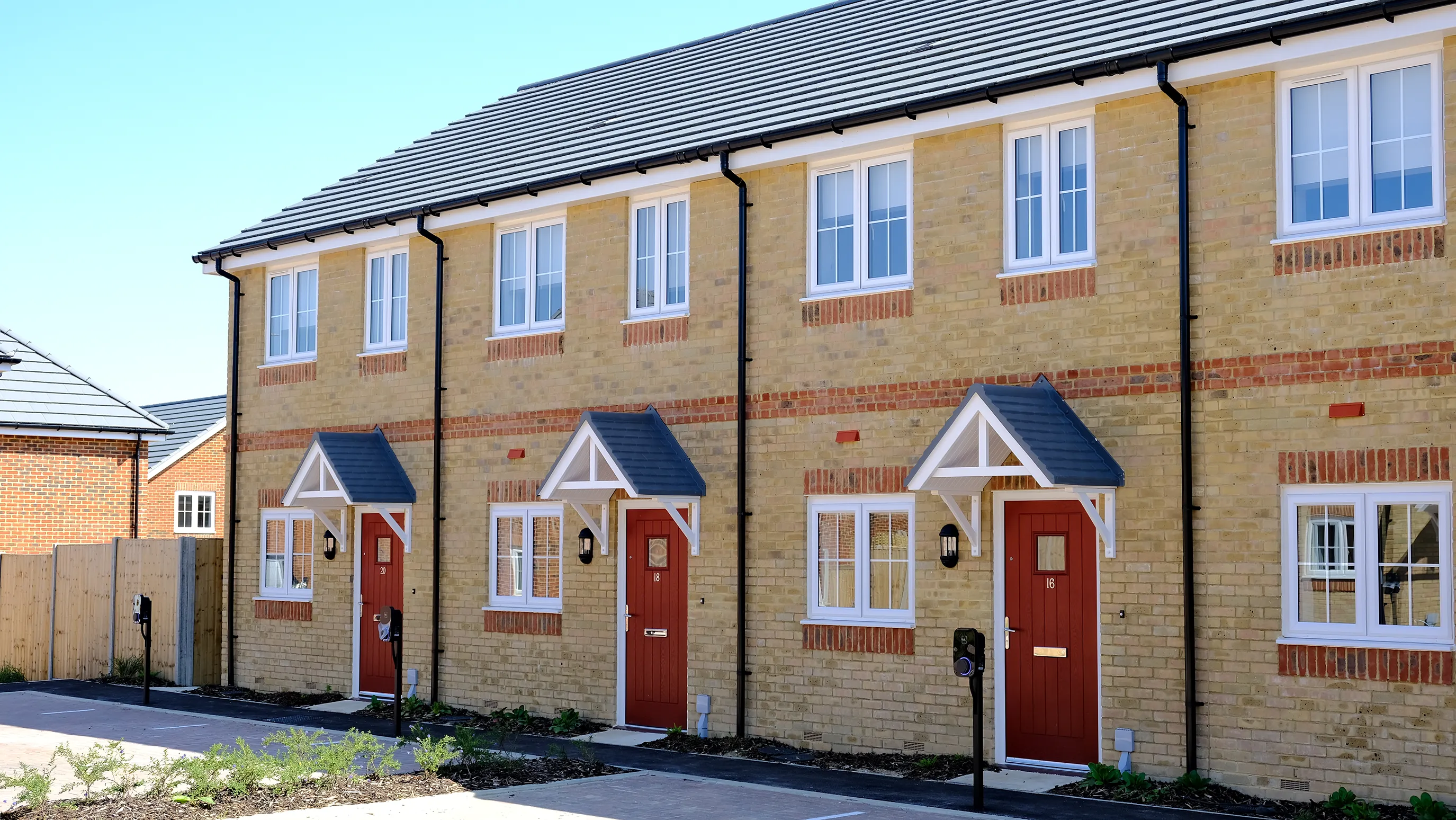 Row of contemporary terraced houses with yellow brick façades, red front doors under small gabled porches, and white-framed windows—lined along a paved walkway with minimal landscaping, reflecting modern suburban housing design.