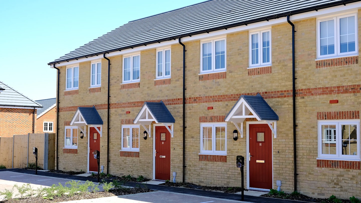 Row of contemporary terraced houses with yellow brick façades, red front doors under small gabled porches, and white-framed windows—lined along a paved walkway with minimal landscaping, reflecting modern suburban housing design.