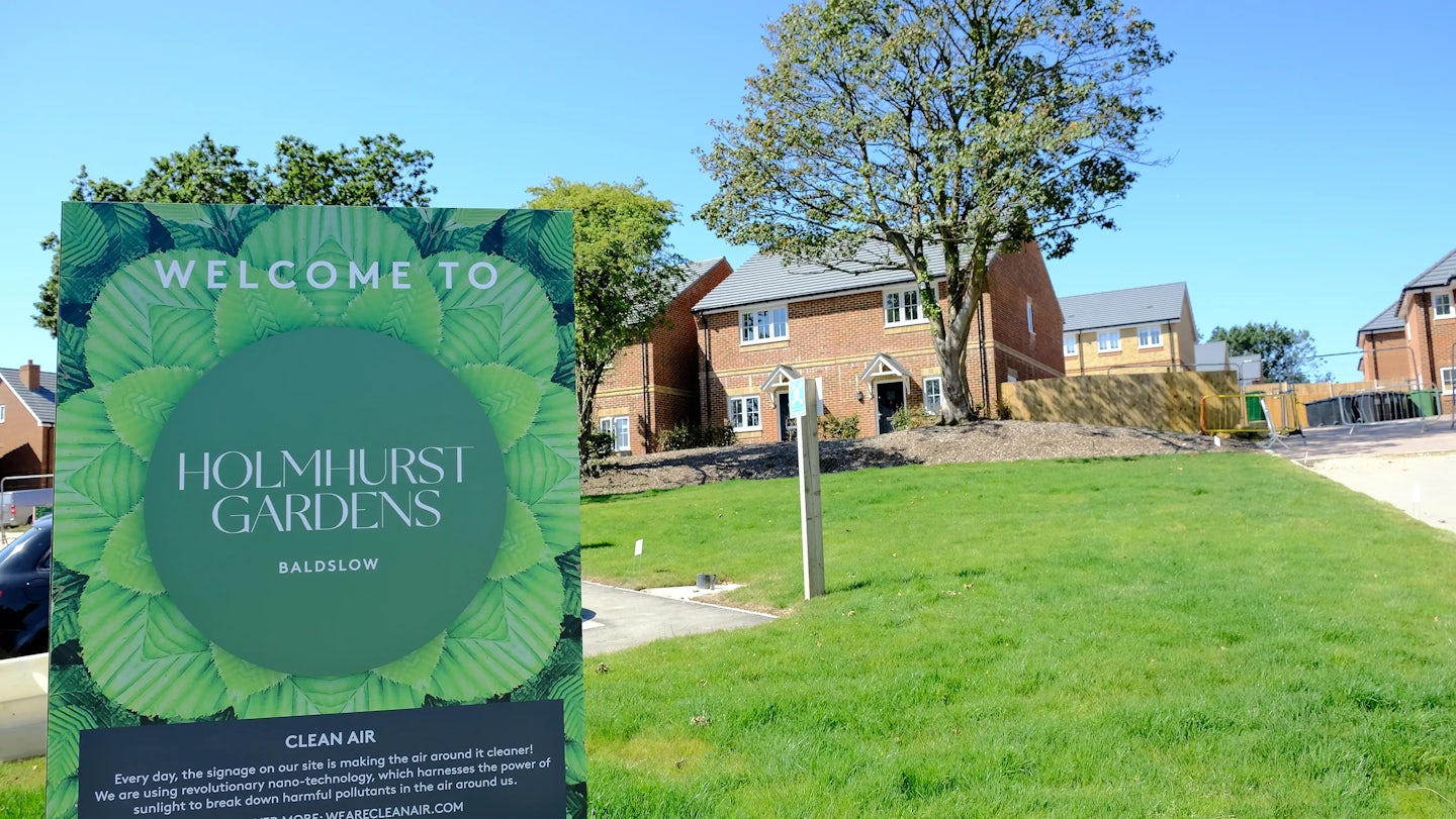 Sunny residential scene with new red-brick houses, a green lawn, and a large roadside sign reading 'Welcome to Holmhurst Gardens, Baldslow,' adorned with leafy graphics—conveying arrival and community identity in a modern housing development.