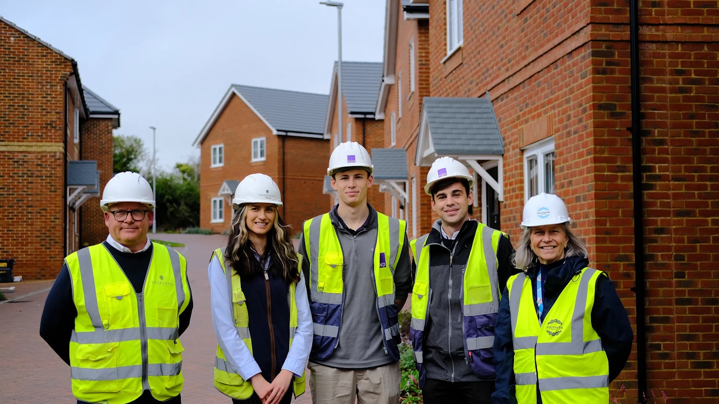 A group of five construction professionals—wearing high-visibility vests and hard hats—stands in front of newly built brick houses on a residential street. Their stance suggests collaboration or site inspection, with the row of red-brick homes, pitched grey roofs, and crisp white-framed windows forming a clean architectural backdrop. The scene evokes themes of teamwork, housing development, and community-building.