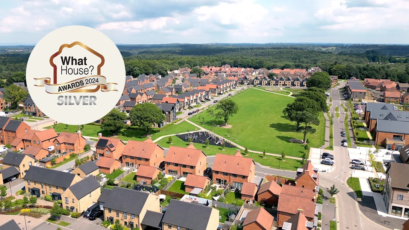 Aerial view of a well-planned residential neighborhood, featuring neat rows of detached and semi-detached homes with red and beige roofs. A large, grassy park anchors the center of the scene, surrounded by tree-lined streets and walking paths. In the top-left corner, a circular badge reads “What House? Awards 2024 Silver,” suggesting the development has earned recognition for design or quality.