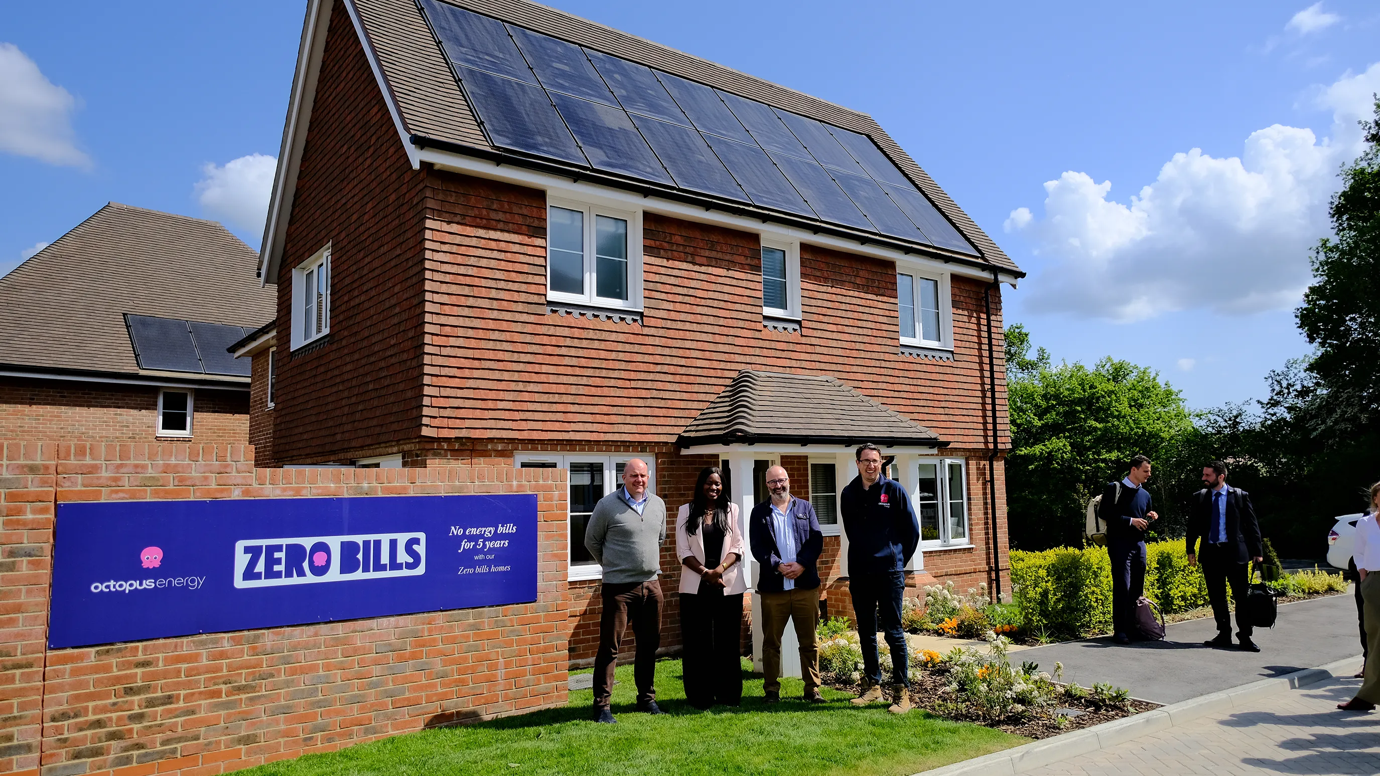 Group of people standing outside a brick house with rooftop solar panels and a sign reading “Octopus Energy ZERO BILLS No energy bills for 5 years.” The house is surrounded by greenery under a partly cloudy sky, suggesting a sustainable, community-focused housing initiative