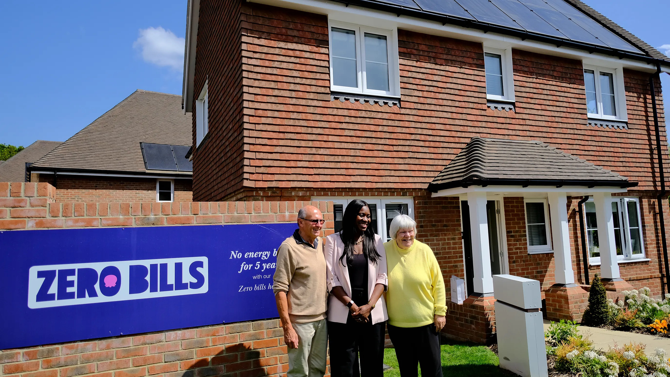 Three people stand proudly in front of a red-brick house with rooftop solar panels. A sign on the wall reads ‘ZERO BILLS – No energy bills for 5 years with our Zero bills home,’ highlighting the home’s eco-friendly promise and financial incentive. The mood is hopeful and forward-looking, reinforcing themes of sustainability and community innovation.