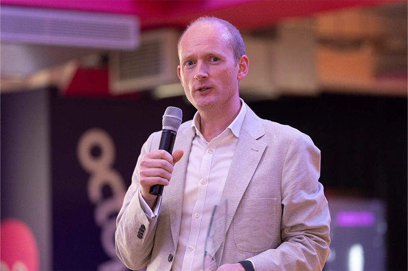 A person in a light blazer and white shirt speaks confidently into a microphone on a stage. Behind them, a dark wall features a white abstract line design with red accents, suggesting a branded or formal event setting. The moment captures poised public speaking and professional engagement.
