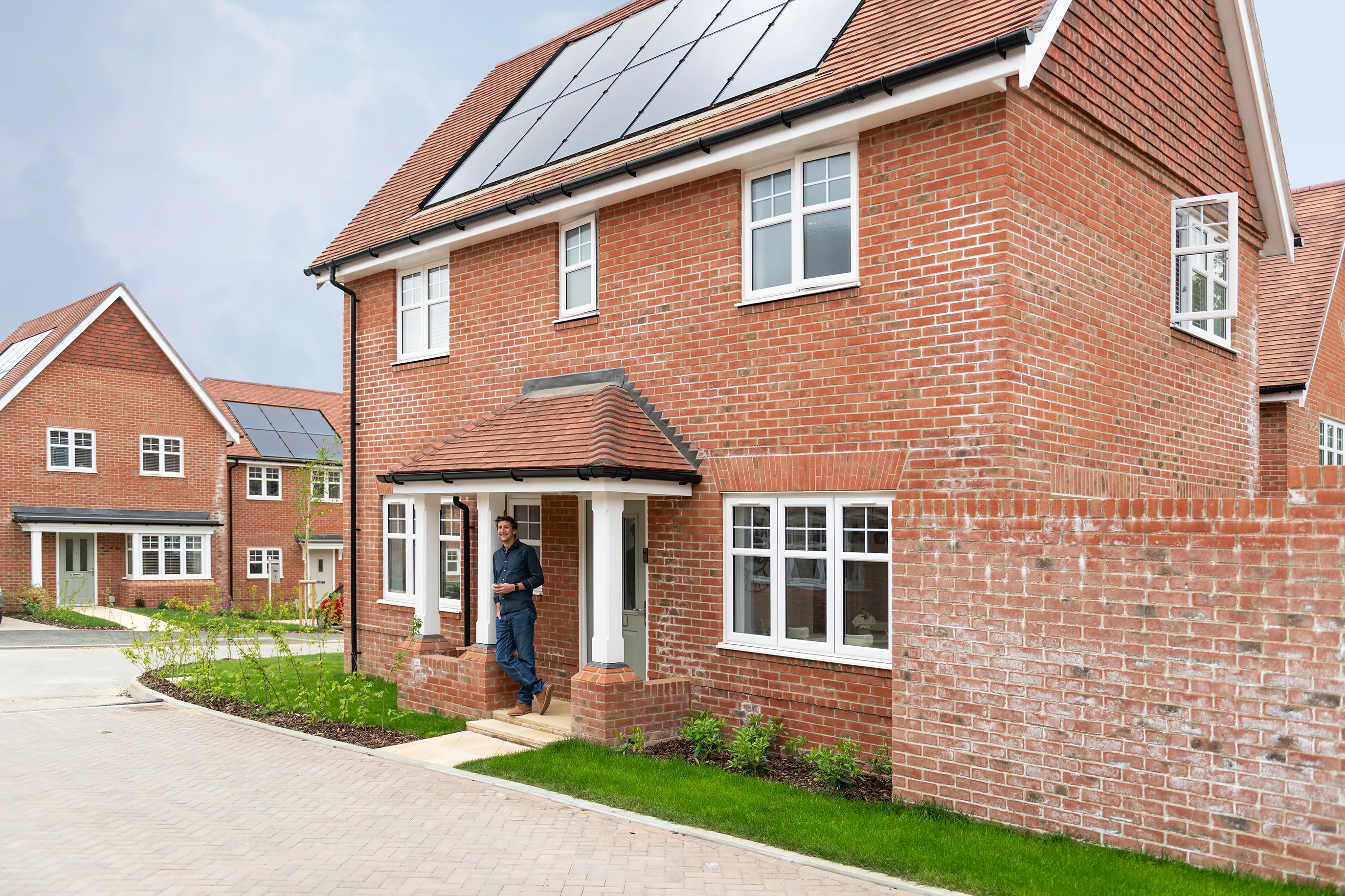 A person stands at the entrance of a modern red-brick house with rooftop solar panels. The front yard features neatly trimmed grass and small plants, suggesting a well-maintained suburban setting. Similar homes appear in the background, reinforcing a community focused on eco-conscious living.