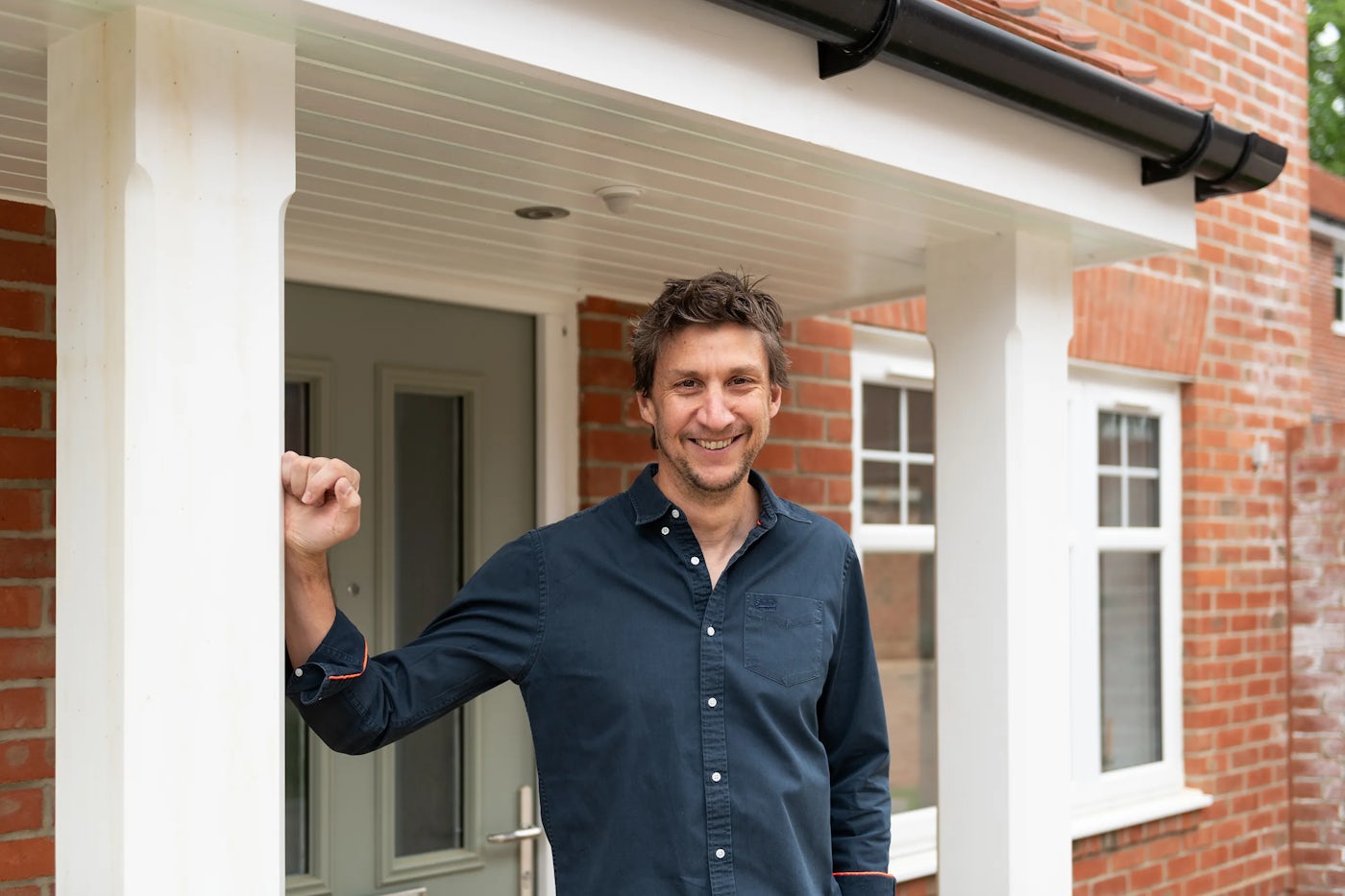 A person in a navy button-up shirt leans casually against a white column on the porch of a red-brick house. Their right arm rests overhead on the column, and they face the camera with a composed expression. Behind them, a grey front door with glass panels and silver hardware is flanked by white-framed windows, conveying a tidy residential facade.