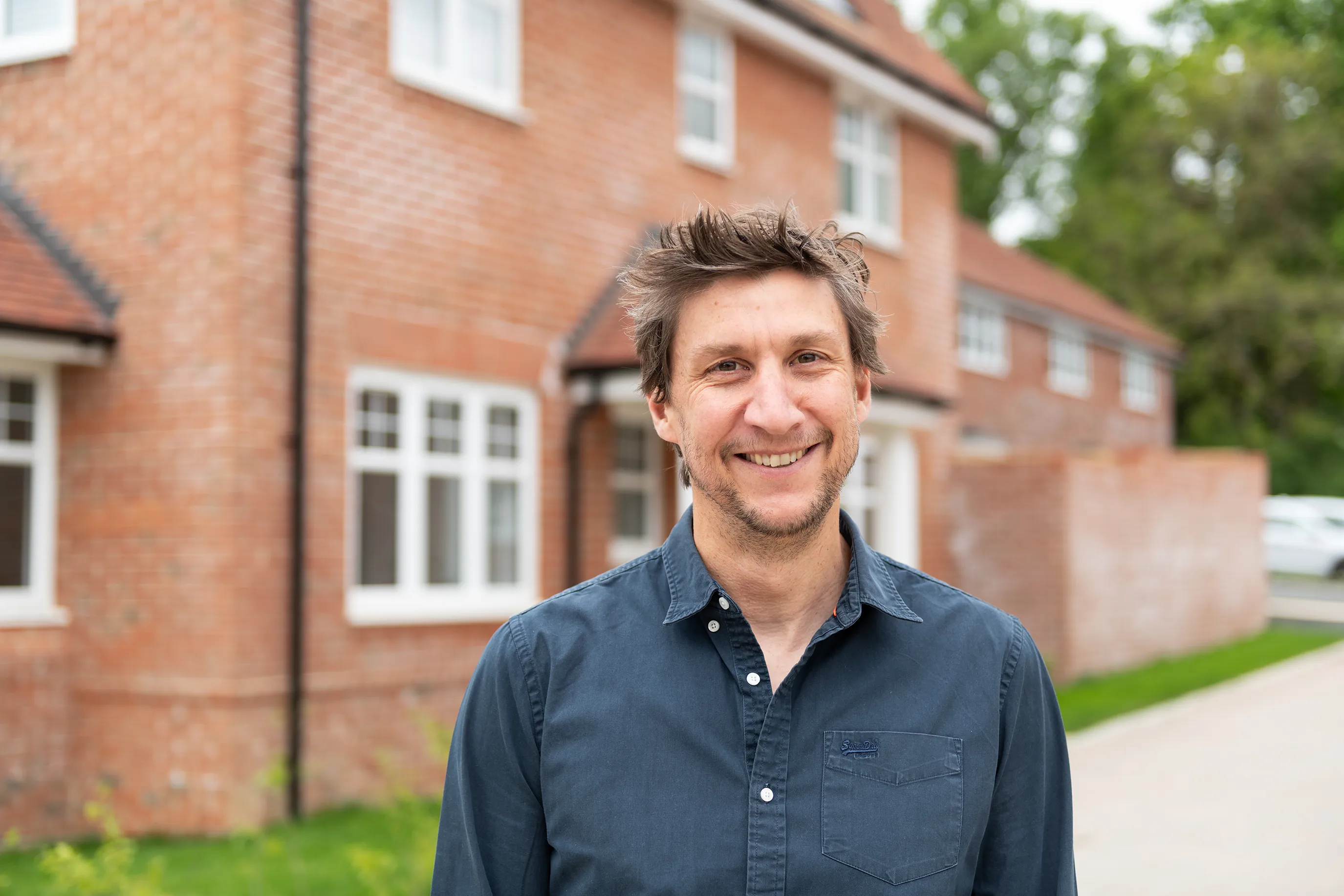 A person stands in a driveway in front of a red-brick home with white-framed windows and a pitched roof. Dressed in a navy button-up shirt, they face the camera with a relaxed posture. Another similar house and lush greenery surround the scene, evoking a quiet, suburban atmosphere.