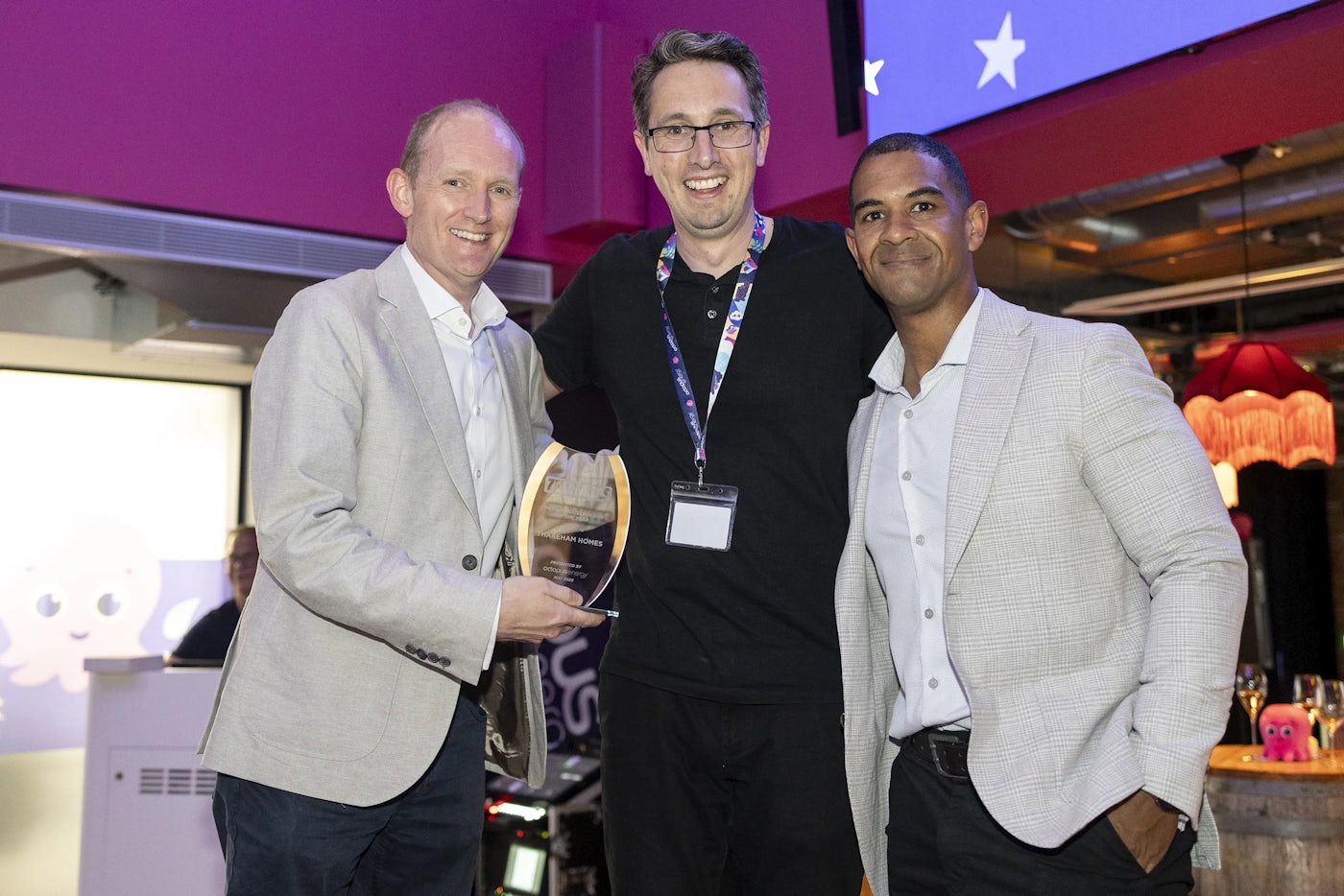 Three individuals stand together in a celebratory moment, one holding a transparent award plaque that reads “American Homes.” Dressed in business attire and event badges, they pose against a pink-toned wall with exposed ceiling fixtures, suggesting a formal yet modern setting. The scene conveys achievement, collaboration, and industry recognition.