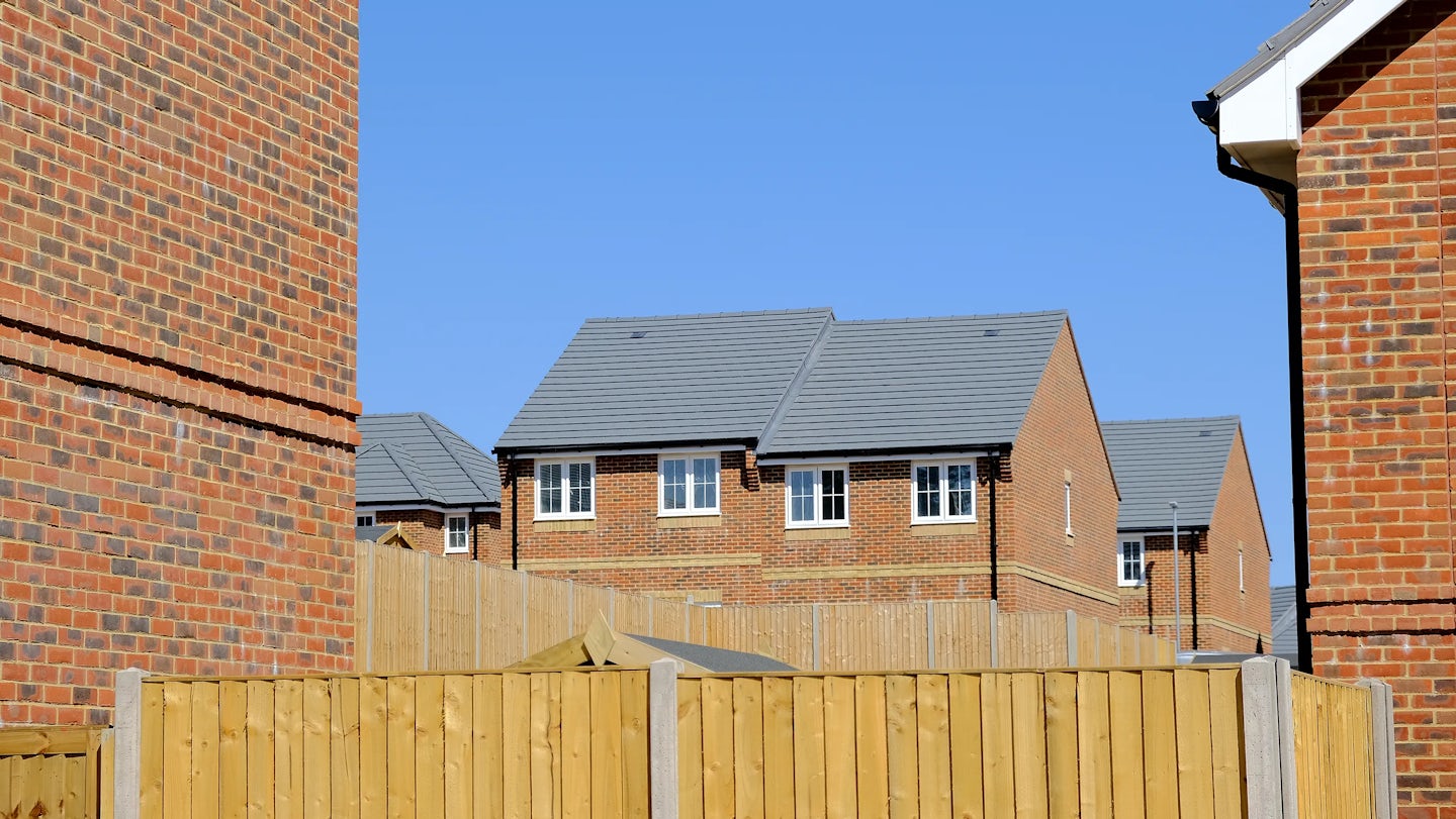 Row of brick houses with grey gabled roofs and white-framed windows, set against a bright blue sky. Wooden fences divide the properties, and each home has a small paved path leading to the entrance. The scene captures a clean, orderly suburban street on a sunny day.
