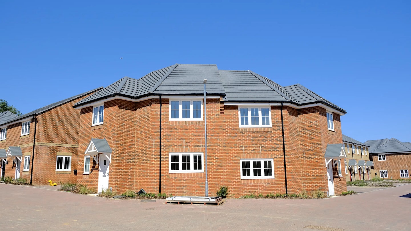 Row of contemporary brick houses with grey gabled roofs under a bright blue sky, featuring white-framed windows and doors. A streetlamp stands in front of the central home, and each property has a small paved area or pathway leading to the entrance. The uniform layout and clean lines suggest a modern suburban development.