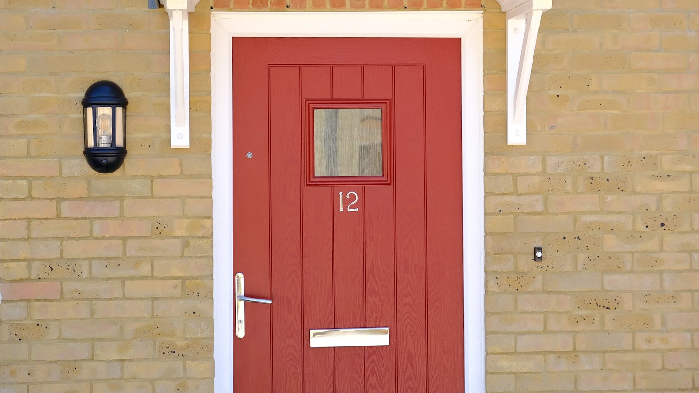 Red front door set in a brick wall, featuring the number 12 in a circular plaque, a square window near the top, silver hardware including a letterbox and handle, and white trim with ornate corbels supporting a small overhang. A black wall light sits to the left and a doorbell is positioned on the right.