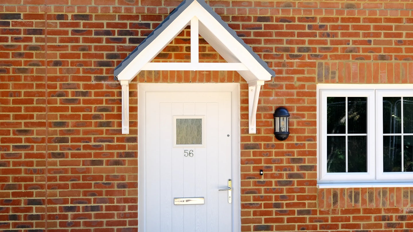 White front door numbered 56 set into a red brick façade, featuring a small rectangular window near the top, a silver letterbox, and a door handle with lock. A small white canopy with supporting brackets shelters the entrance. To the right, a black outdoor light is mounted on the wall beside a white-framed window.
