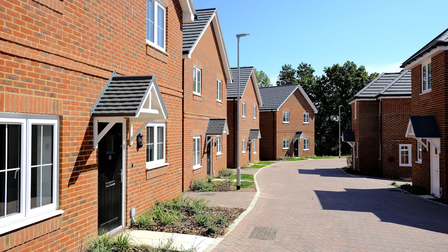 Row of modern brick houses with white-framed windows and black front doors, each sheltered by a small gabled porch. The street is lined with streetlamps and neat patches of greenery, and the scene is set under a clear, sunny blue sky—evoking a calm, well-kept suburban atmosphere.