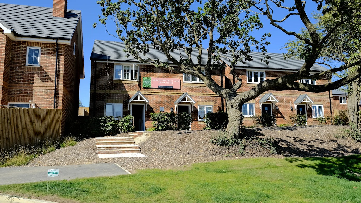 Modern brick houses set behind a landscaped front garden with mulch, greenery, and a large tree casting partial shade. White-framed windows and doors punctuate the façade, with one home displaying signs reading ‘SHOW HOME OPEN’ and ‘SALES OFFICE OPEN.’ Decorative triangular gables accent the entrances, and a short set of steps connects the paved path to the homes. The scene is bathed in bright, natural light beneath a clear blue sky.
