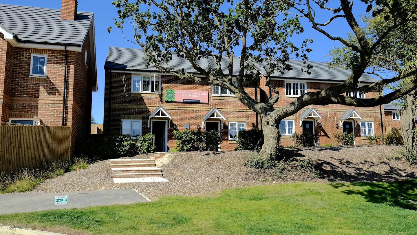 Modern brick houses set behind a landscaped front garden with mulch, greenery, and a large tree casting partial shade. White-framed windows and doors punctuate the façade, with one home displaying signs reading ‘SHOW HOME OPEN’ and ‘SALES OFFICE OPEN.’ Decorative triangular gables accent the entrances, and a short set of steps connects the paved path to the homes. The scene is bathed in bright, natural light beneath a clear blue sky.
