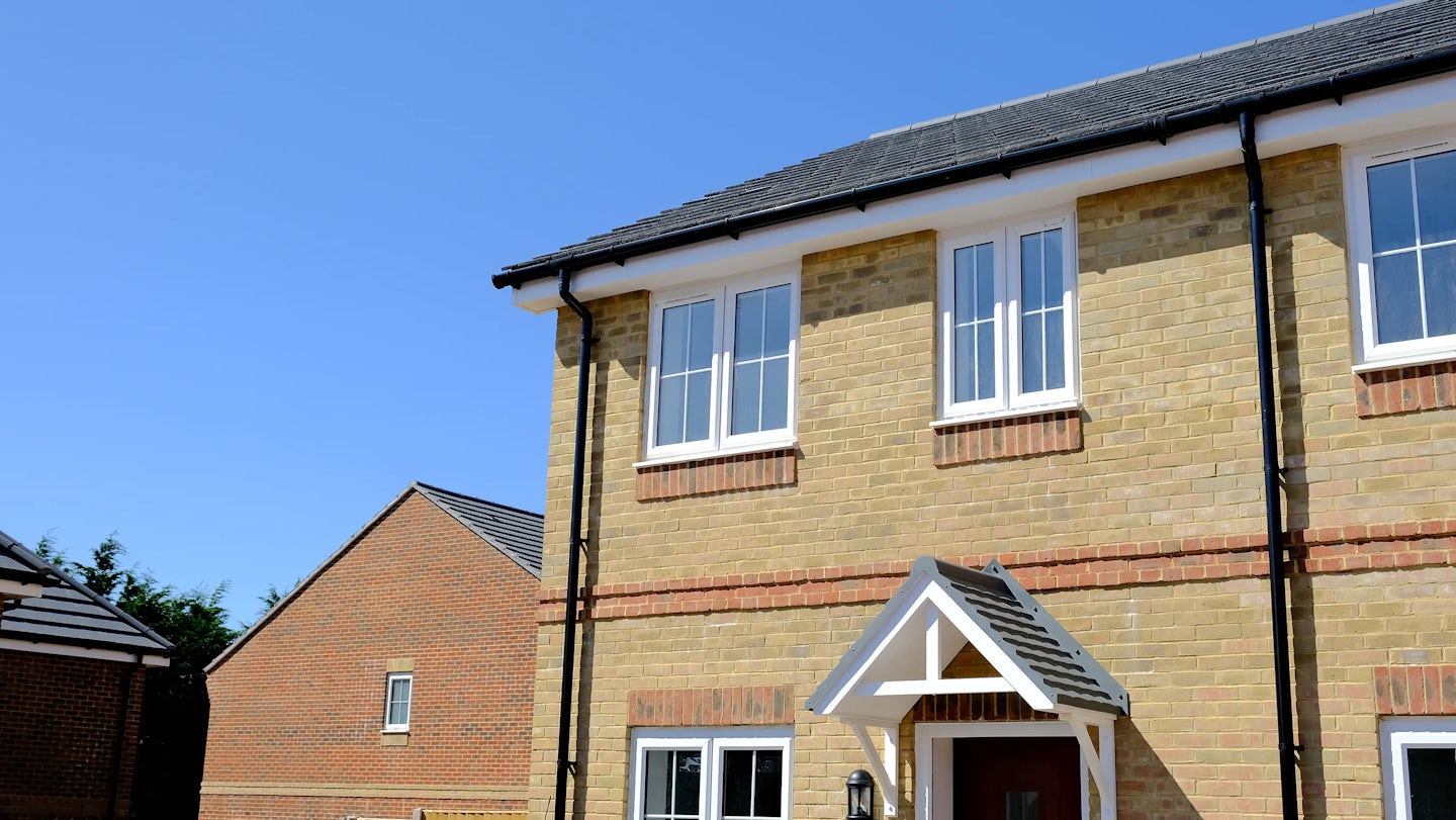 Two-story brick house with a gabled roof and white-framed windows, featuring a small front porch with a triangular canopy over the entrance. Black downspouts run along the front facade, and a clear blue sky brightens the backdrop. Another similar house appears partially in view to the left.