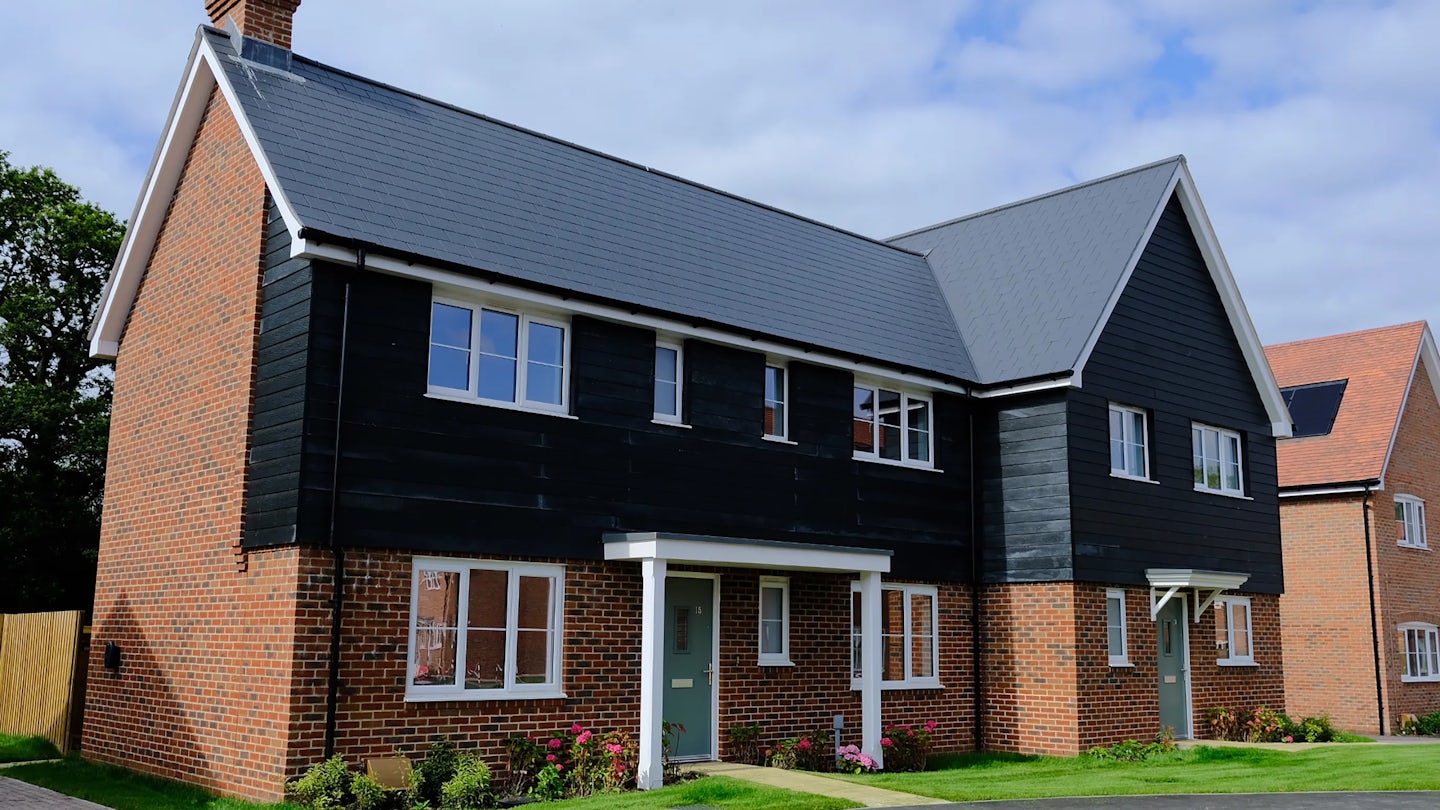 Contemporary two-story house with a mix of red brick and black timber-style siding. The grey shingled roof features a steep pitch with a gable above a bay window on the ground floor. A green front door with a covered porch sits beside flower beds and a manicured lawn. Other similar houses appear in the background beneath a partly cloudy sky.