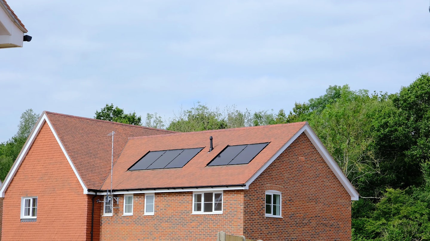 A brick house with a red-tiled roof and two solar panels installed near a satellite dish. The building is surrounded by trees and shrubs, with a partly cloudy sky above—evoking a balance of traditional architecture and eco-conscious living.