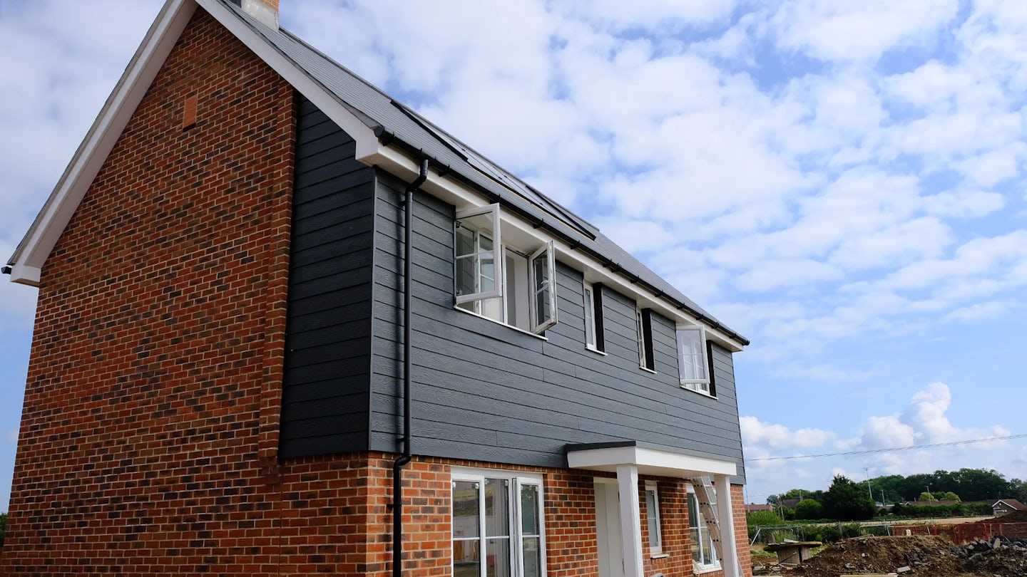Modern two-story house with a mix of brick and dark grey siding, featuring a pitched roof with solar panels. One window is open, revealing a fan inside, and construction materials are visible near the base. The sky is partly cloudy, and trees add greenery to the background.