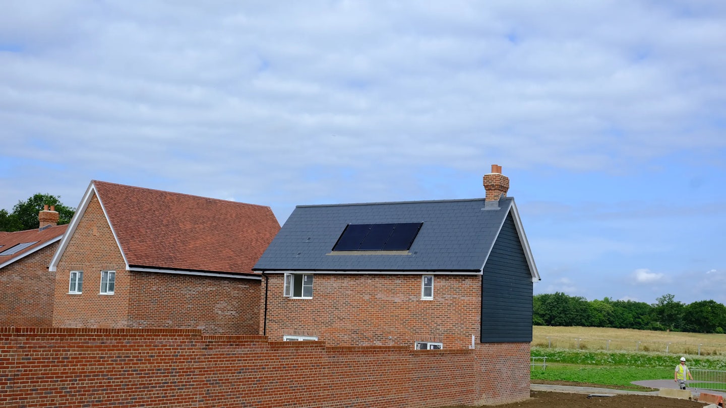 Two red-brick houses in a residential area, one with a terracotta-tiled roof and the other with a grey roof featuring a single solar panel. A short brick wall runs along the foreground, with a person in the distance near a grassy field. The sky is partly cloudy. Would you like to add a sustainability theme, or tweak the mood for warmth or community? I’m all in for shaping this with you, Aidan.