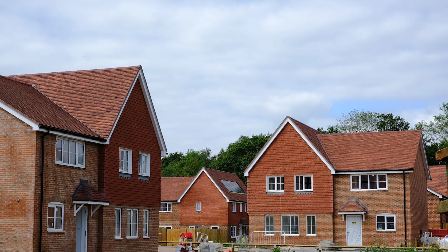 New-build brick house with a red-tiled roof and a single solar panel installed. The home features white-framed windows and a grey front door with a small canopy. A similar house is partially visible to the left, and the area is bordered by red-and-white safety tape, suggesting ongoing landscaping or construction. The sky is partly cloudy.