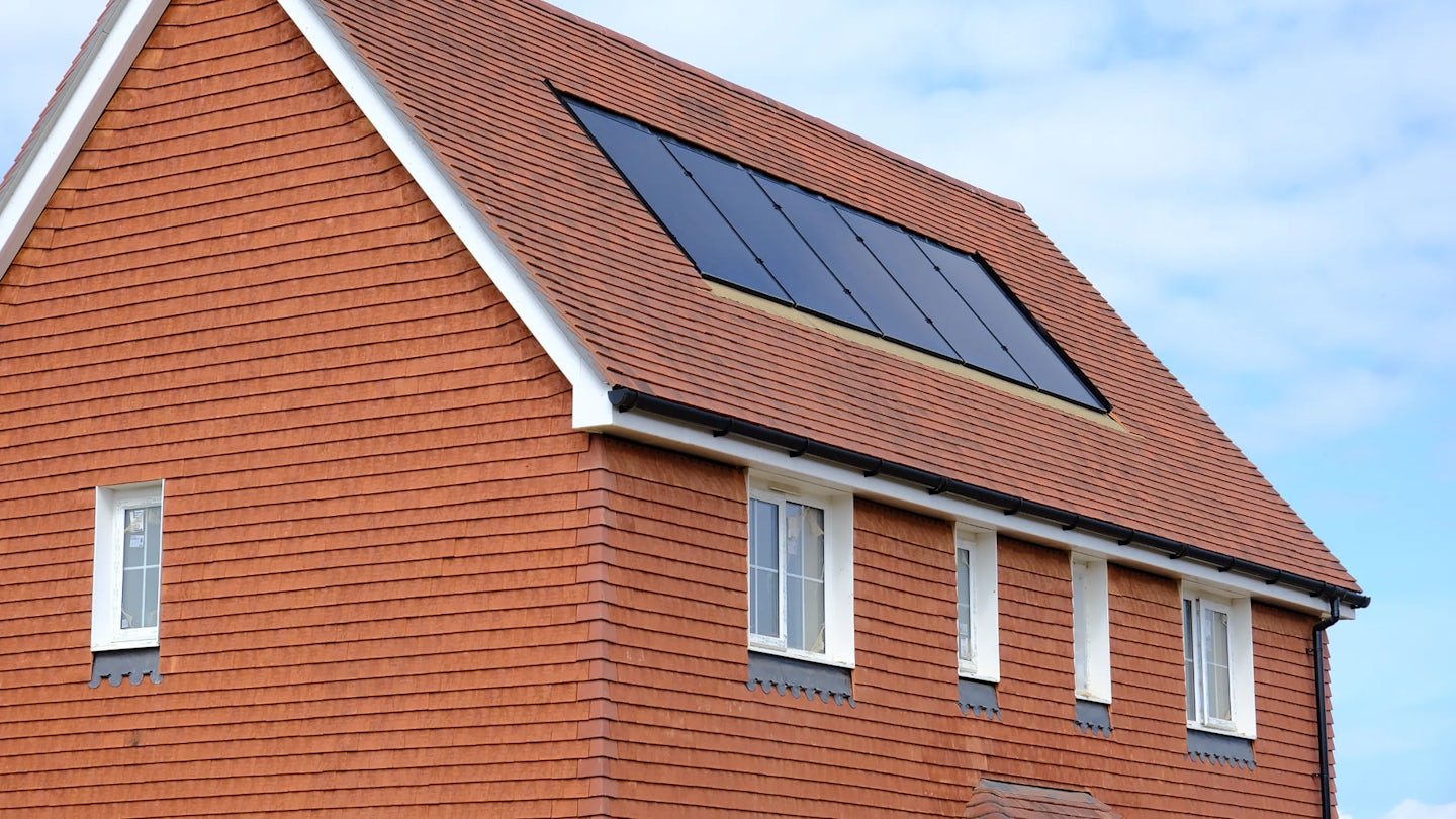A red brick house with a terracotta-tiled roof featuring solar panels, set beneath a partly cloudy sky. Four white-framed windows are evenly spaced on the side of the building, each with a decorative blue panel below