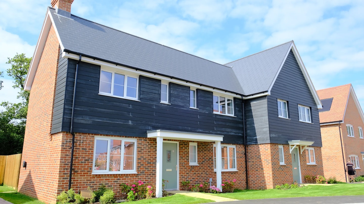 Modern two-story house with a mix of red brick and dark wood siding, featuring a grey-tiled roof and white-framed windows. A tidy front garden with flowering plants and a manicured lawn adds a touch of charm. A similar home appears in the background beneath a clear, lightly clouded sky.