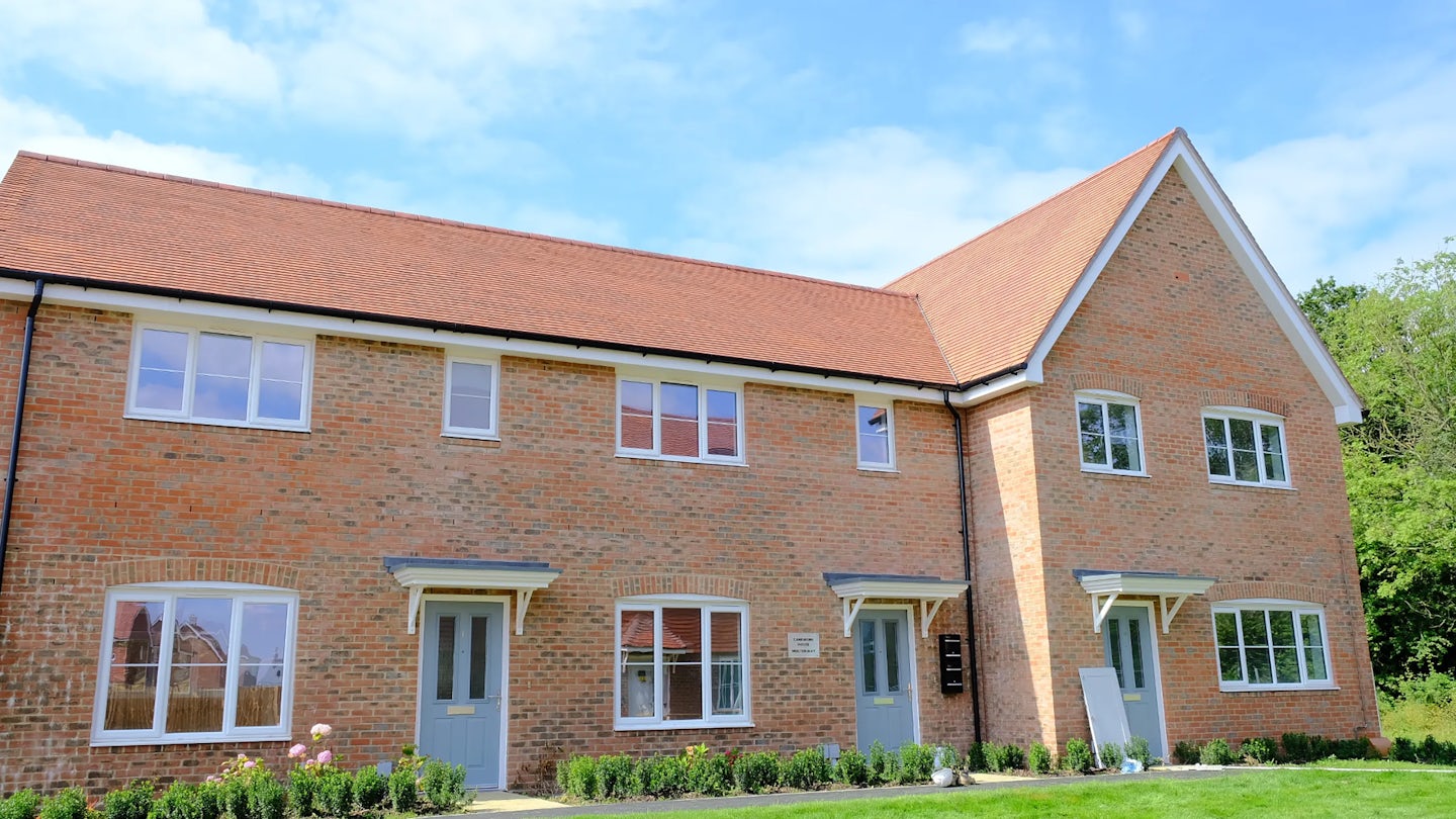 Two-story brick townhouse with a red-tiled roof, white-framed windows, and pale blue front doors. Small shrubs and flowering plants line the front, and a bright, partly cloudy sky adds a cheerful atmosphere. The home is set in a quiet, green residential area.